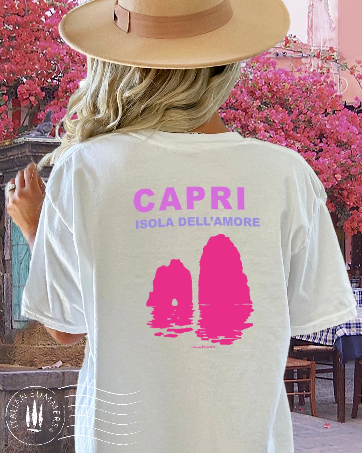 A youg woman stands in front of a quaint Italian island restaurant covered in pink bougainvilleas. She is standing with her back to us wearing a soft white cotton shirt with a pink and fuchsia print depicting the Faraglioni islets of the famous island of Capri. The text on the print states " Capri Isola dell'Amore" Capri, island of love.