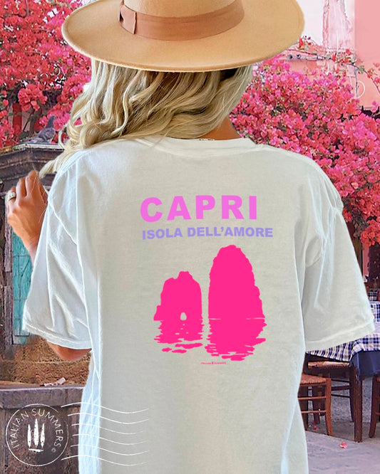 A youg woman stands in front of a quaint Italian island restaurant covered in pink bougainvilleas.  She is standing with her back to us  wearing a soft white cotton shirt with a pink and fuchsia print depicting the Faraglioni islets  of the famous island of Capri.  The text on the print states " Capri Isola dell'Amore" Capri, island of love.