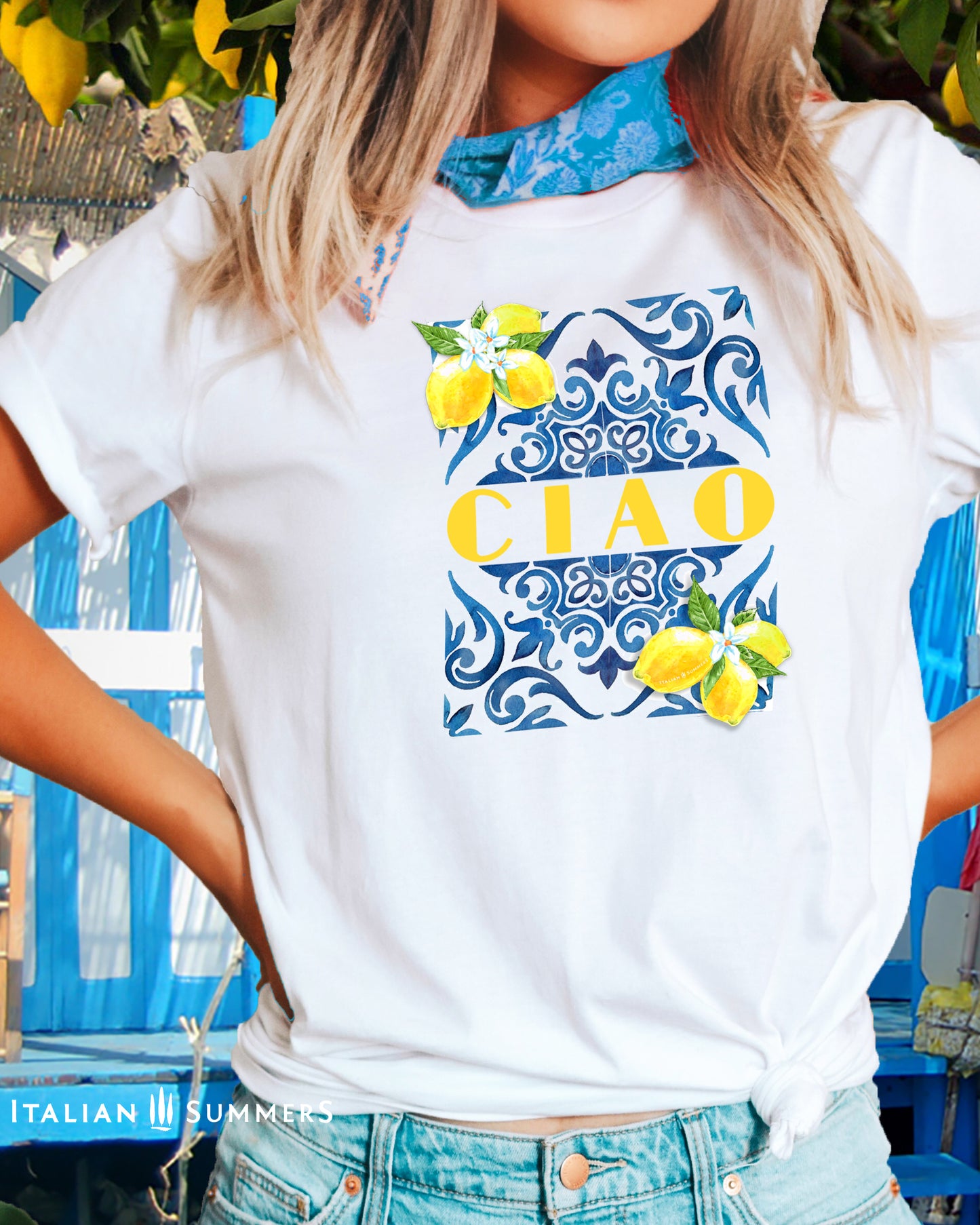 A blonde girl wears a white cotton T shirt with the print of a blue Italian Maiolica tile on the front, framed by lemons with blooms and the word CIAO in large Art Deco yellow characters in the middle of the composition