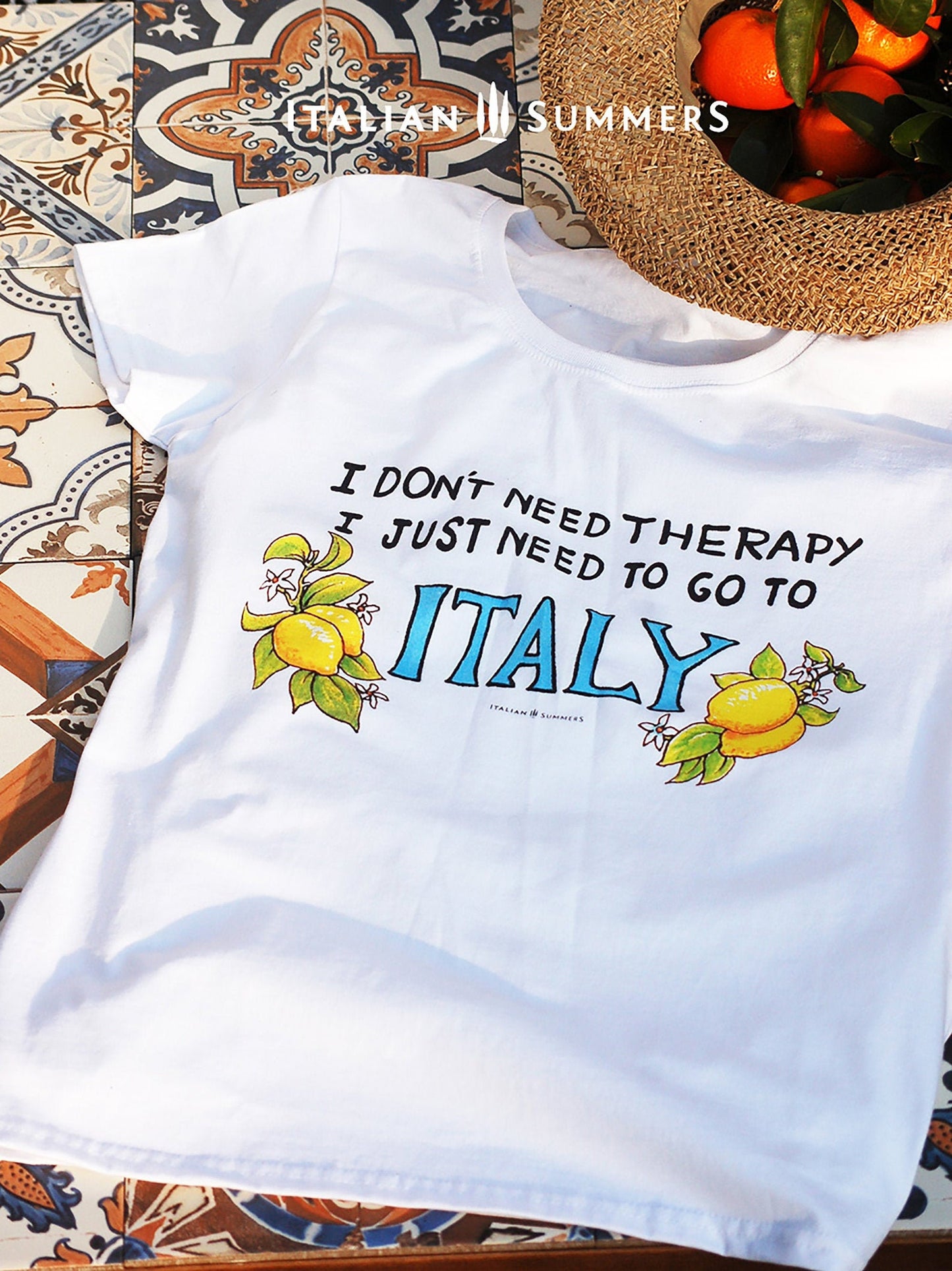 Italy inspired white cotton T shirt with the quote " I don't need therapy I just need to go to Italy" the word " Italy" Is in capital blue letters and flanked by hand-painted bunches of lemons with flowers. A best seller made by Italian Summers