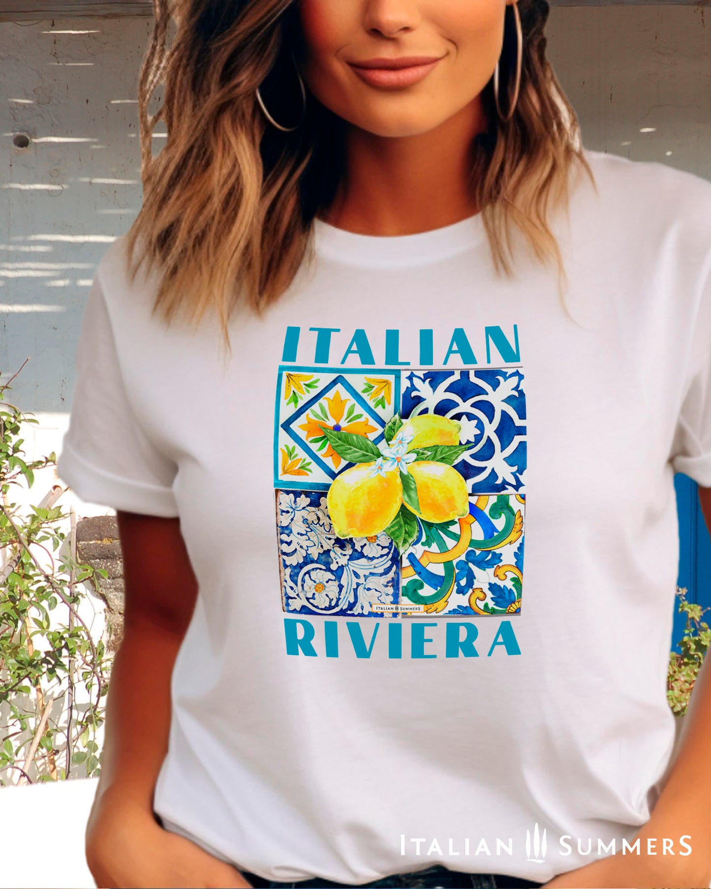 An all cotton T shirt with a Mediterranean tile design on the chest and a large cluster of Amalfi Coast lemons on top of the tiles. The lemons are bright yellow with green leaves and flowers. A blue text states: ITALIAN RIVIERA