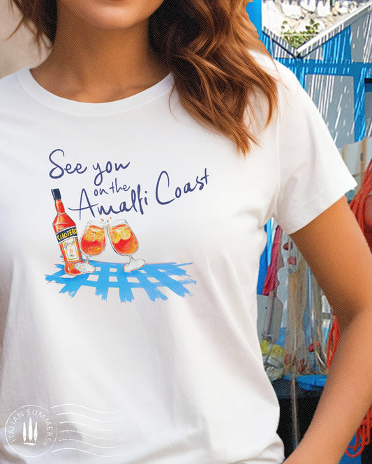 Italy inspired white t-shirt with the text See you on the Amalfi Coast in navy blue and a sketch in water color with an Aperol bottle and 2 glassis filled with Aperol Spritz on a bright blue table cloth.