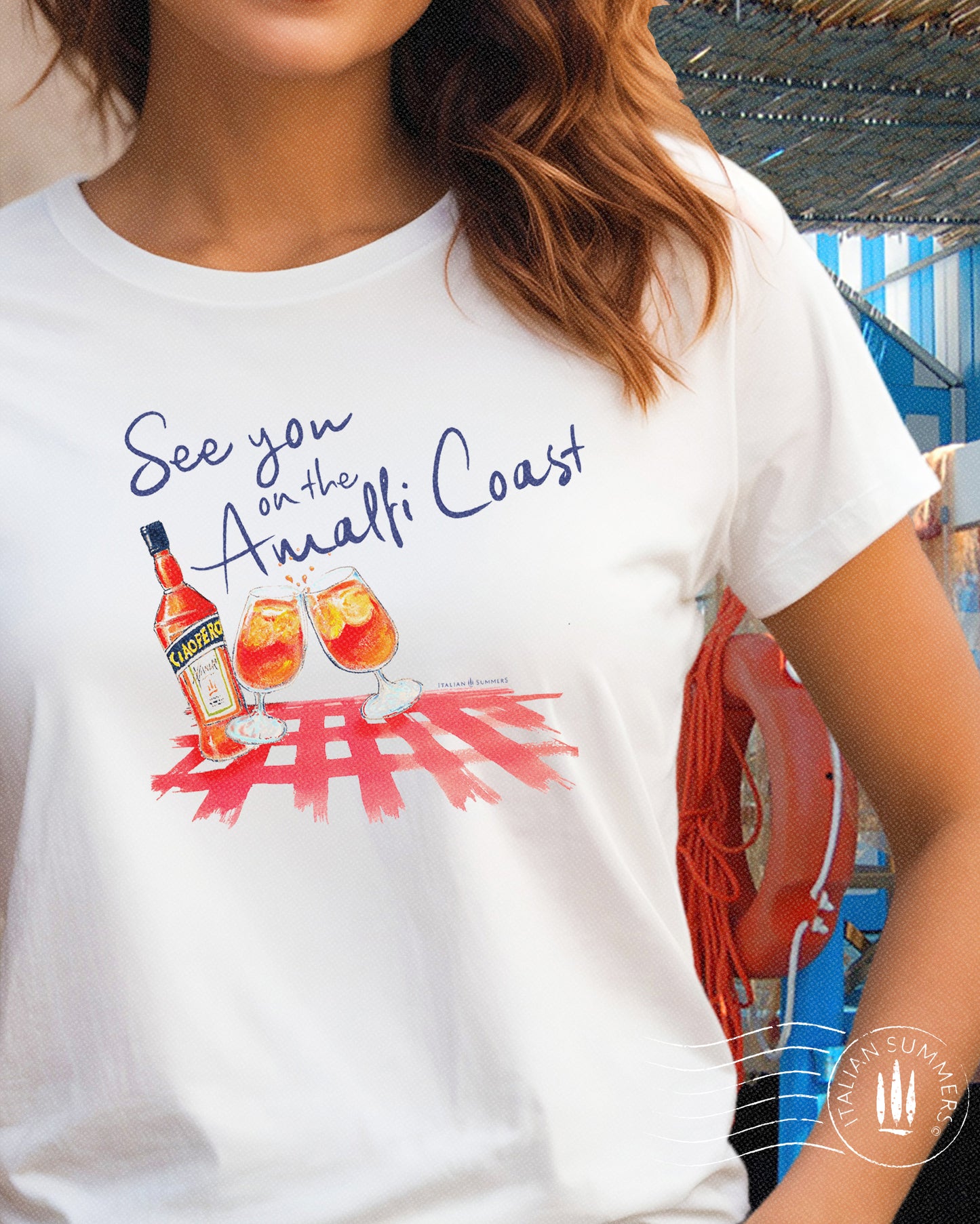 Italy inspired white t-shirt with the text See you on the Amalfi Coast in navy blue and a sketch in water color with an Aperol bottle and 2 glassis filled with Aperol Spritz on a bright red table cloth.
