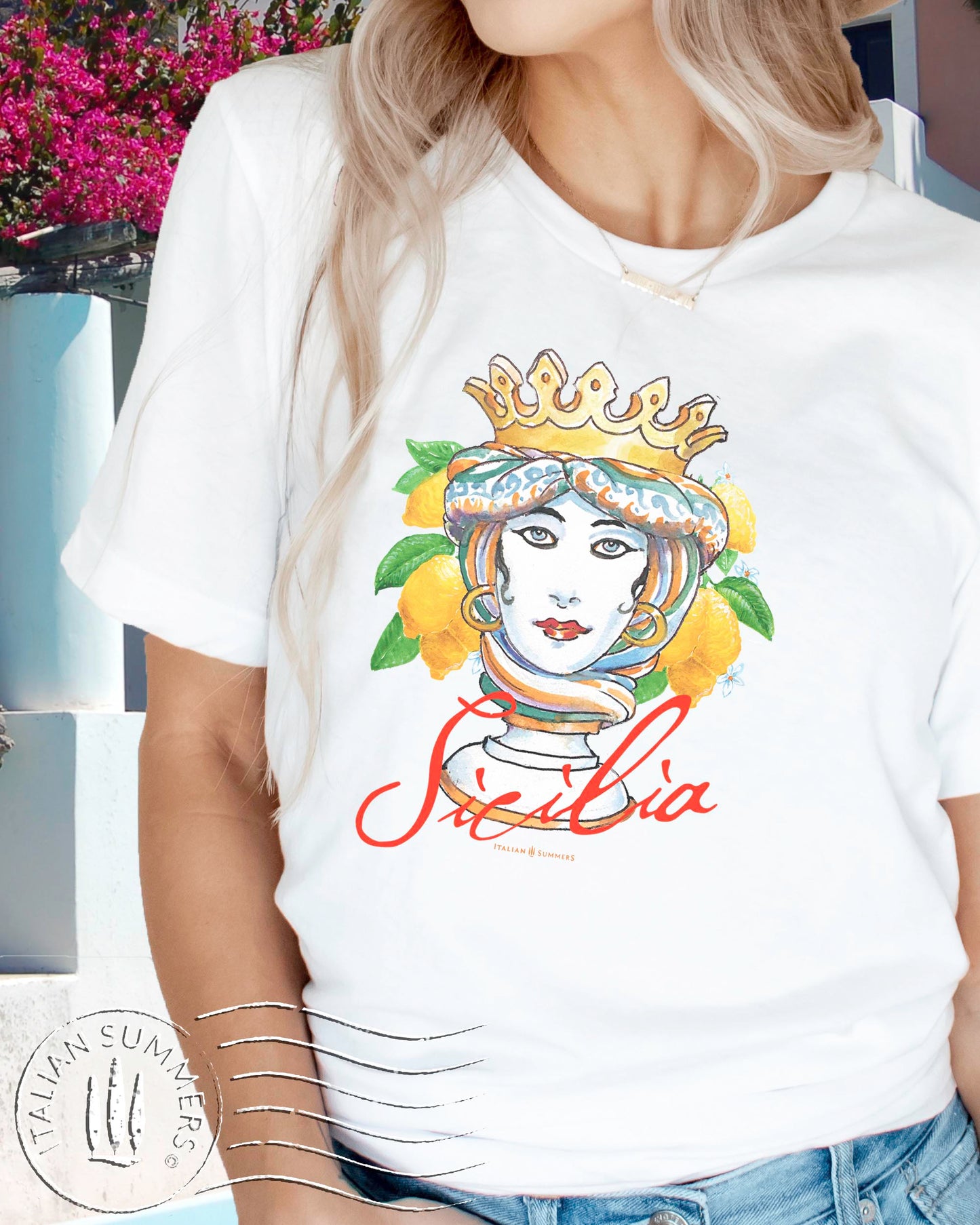 An all cotton white T-shirt featuring a frontal print of a Sicilian Moor's Head of a woman with bright turban and golden crown, surrounded by a vibrant cluster of leafy and flowery Sicilian Lemons. The cursive text : Sicilia is printed un the bottom in red. Reminiscent of the famous Sicilian ceramics from Caltagirone.