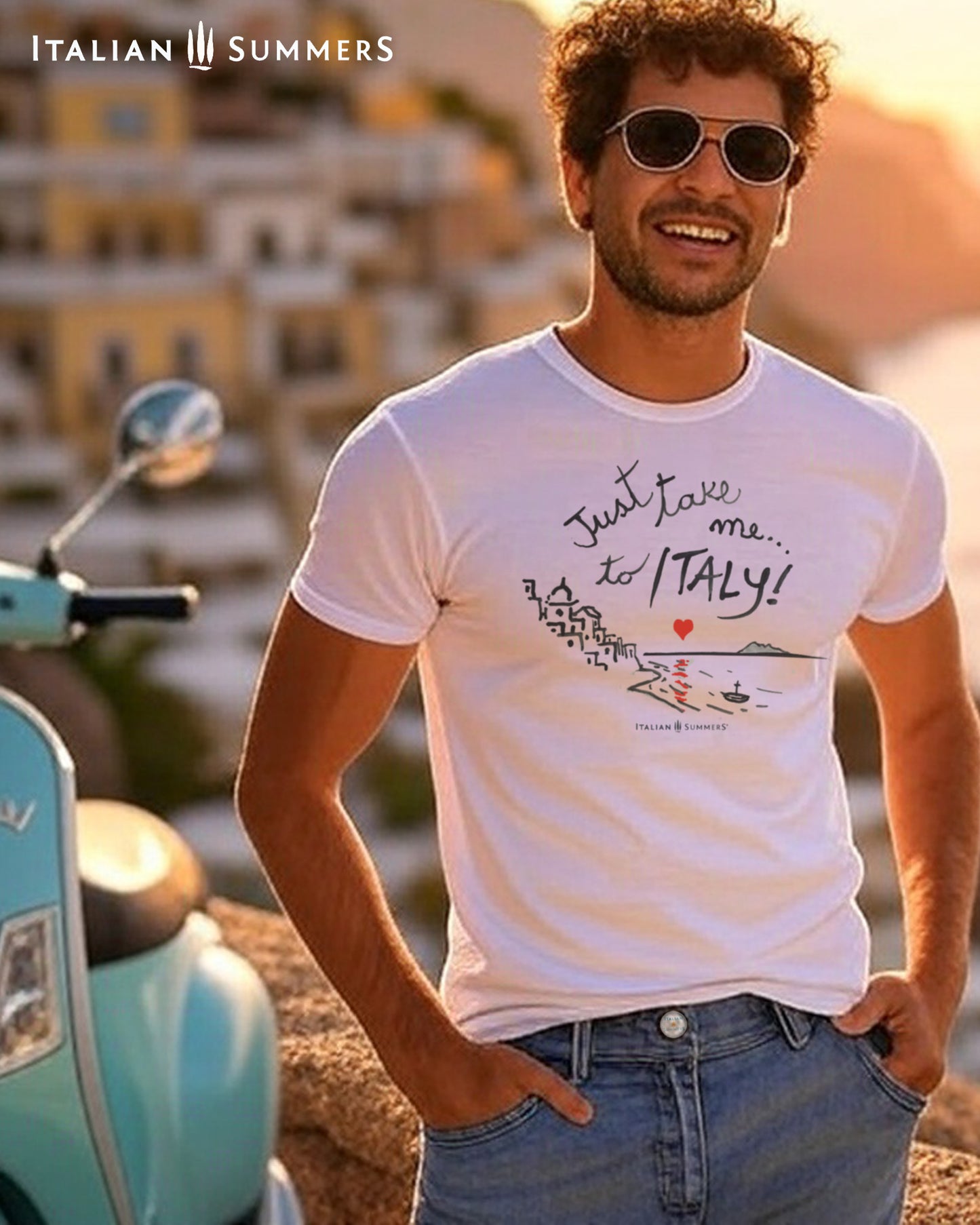 An Italian young man wearing a Italy-inspired Unisex crew neck T shirt with a print of a hand-painted stylized Amalfi Coast village with a red heart setting on the sea and reflecting its glow on the waters. The hand-painted phrase "Just take me to Italy" is above the composition