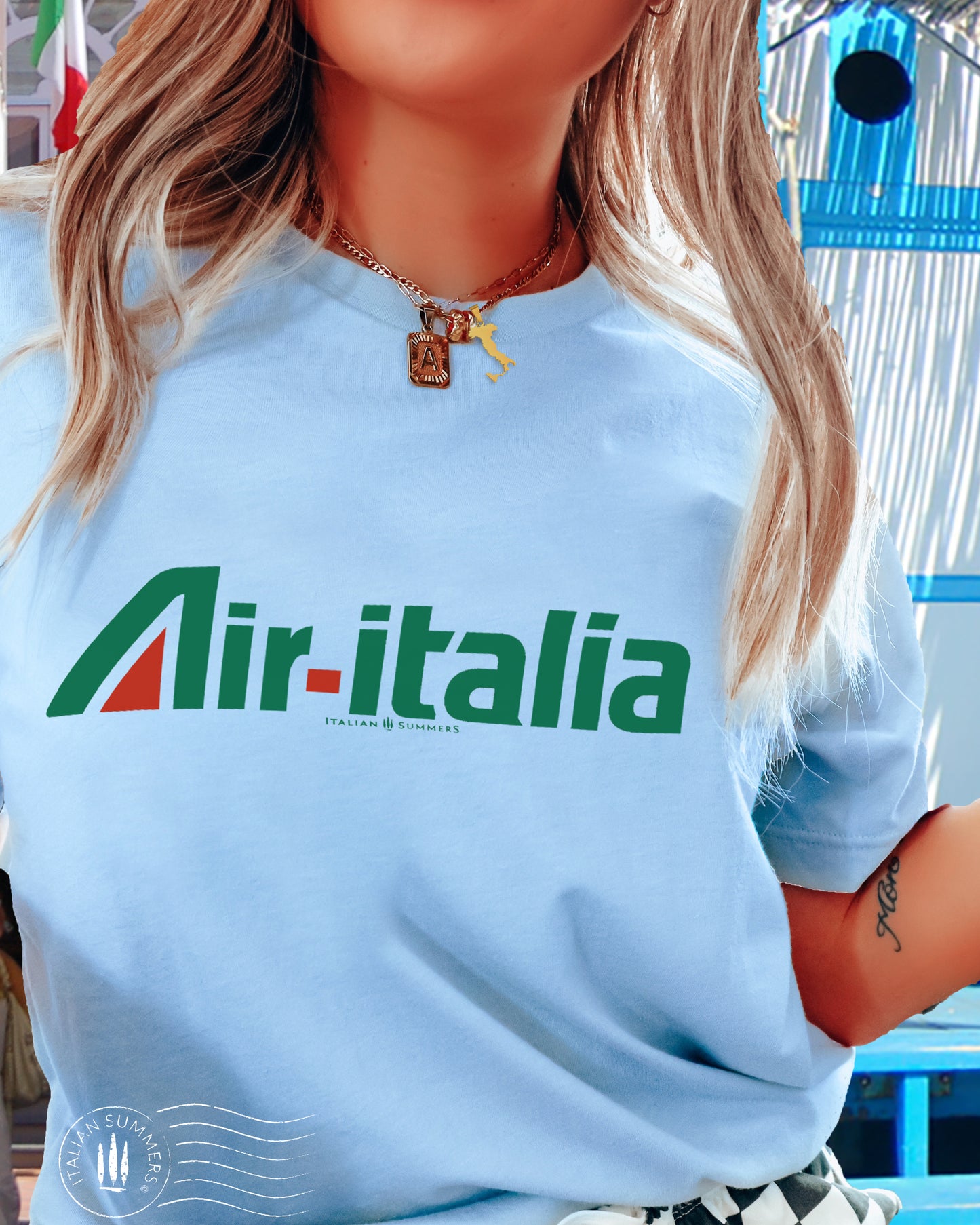 A girl is wearing an all cotton light blue T-shirt with a large green and red Logo of Air-Italia airlines. A retro design of the nostalgic 1960s in Italy when Alitalia was the national airline. Sure to bring memories to Italy aficionados