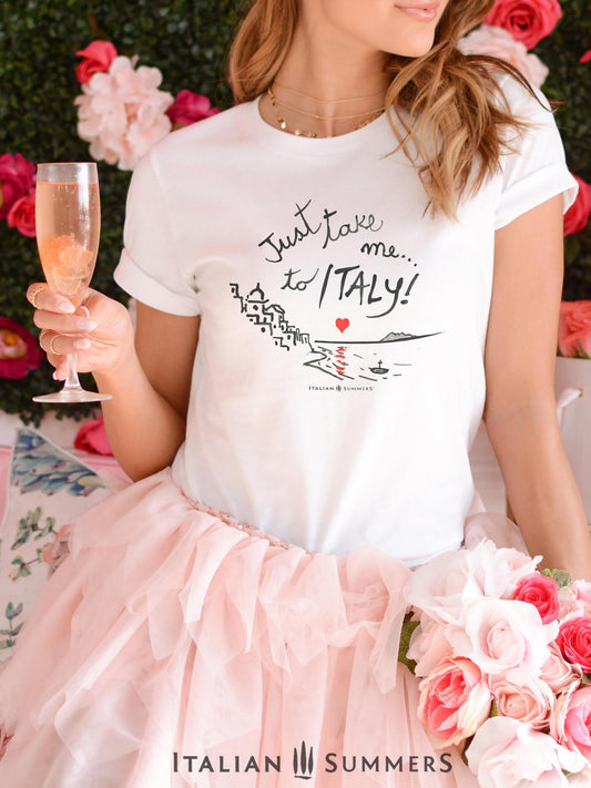 A girl, likely a bachelorette with a fluffy pink tutu skirt is wearing a lItaly-inspired Unisex crew neck T shirt with a print of a hand-painted  stylized Amalfi Coast village with a red heart setting on the sea and reflecting its glow on the waters. The hand-painted phrase "Just take me to Italy" is above the composition