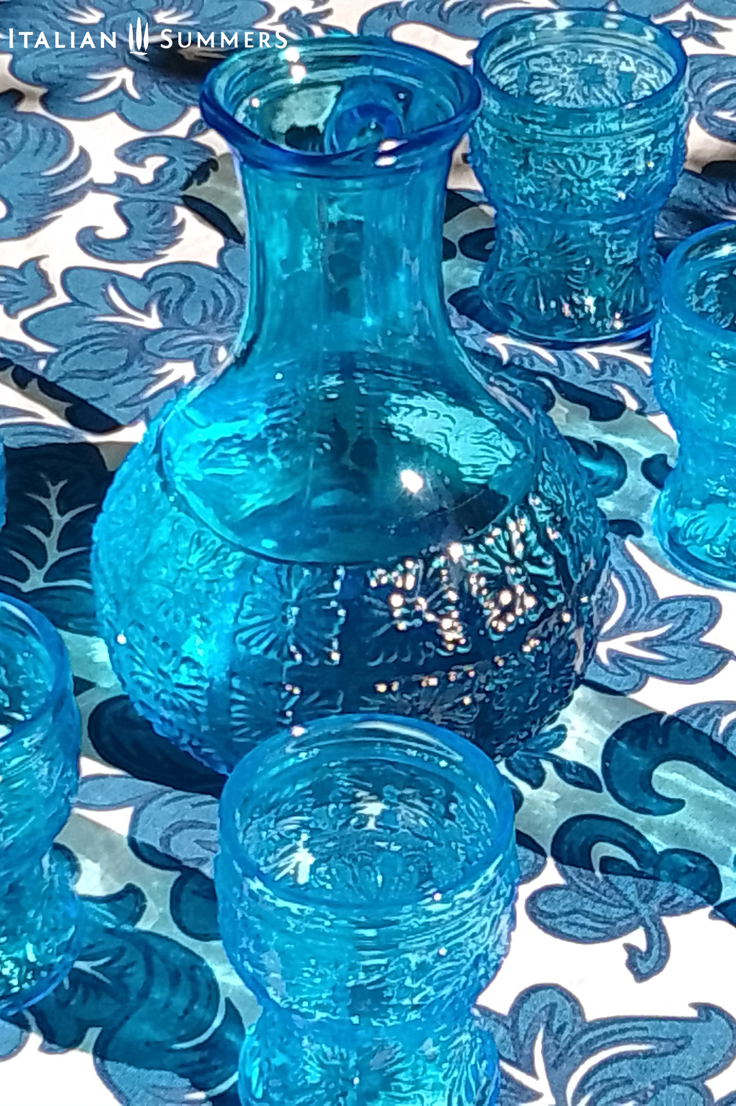 A blue glass carafe and six tumblers set, all embossed in flower patterns from the 1970s Italy, sits on a table with a colorful and bright tablecloth with blue floral patterns.