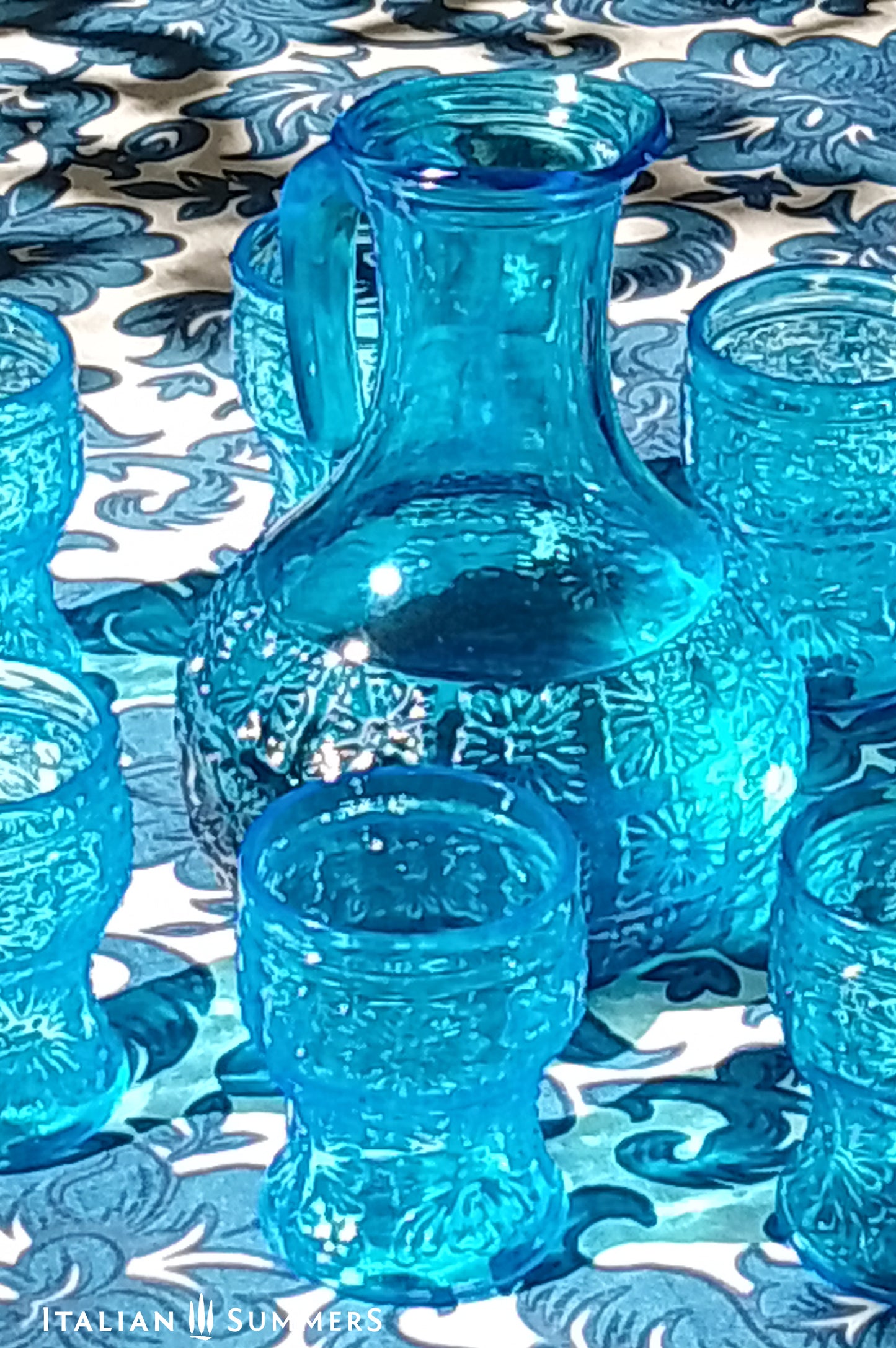 A blue glass carafe and six tumblers set, all embossed in flower patterns from the 1970s Italy, sits on a table with a colorful and bright tablecloth with blue floral patterns.