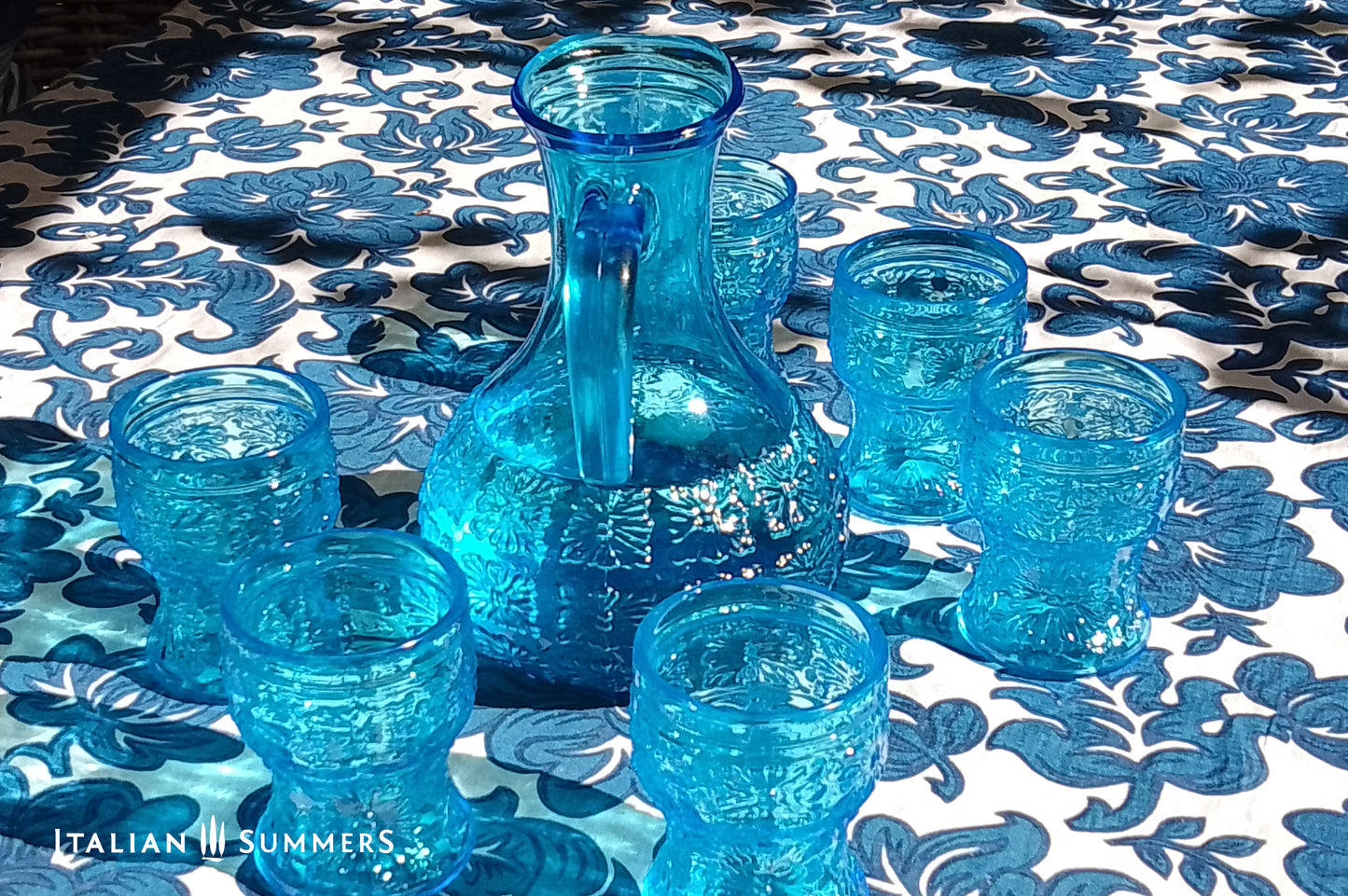 A blue glass carafe and six tumblers set, all embossed in flower patterns from the 1970s Italy, sits on a table with a colorful and bright tablecloth with blue floral patterns.