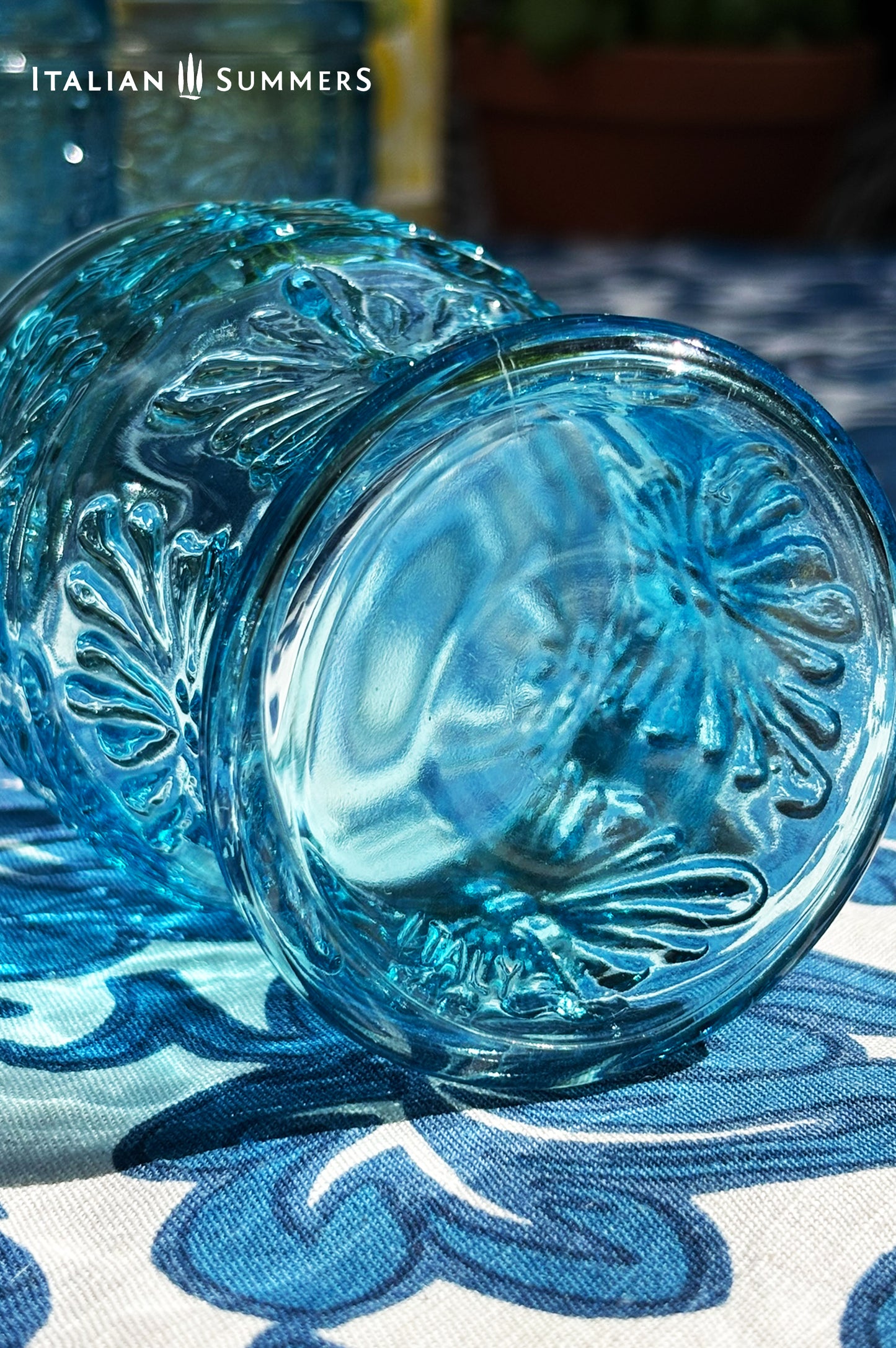 A blue glass carafe and six tumblers set, all embossed in flower patterns from the 1970s Italy, sits on a table with a colorful and bright tablecloth with blue floral patterns.