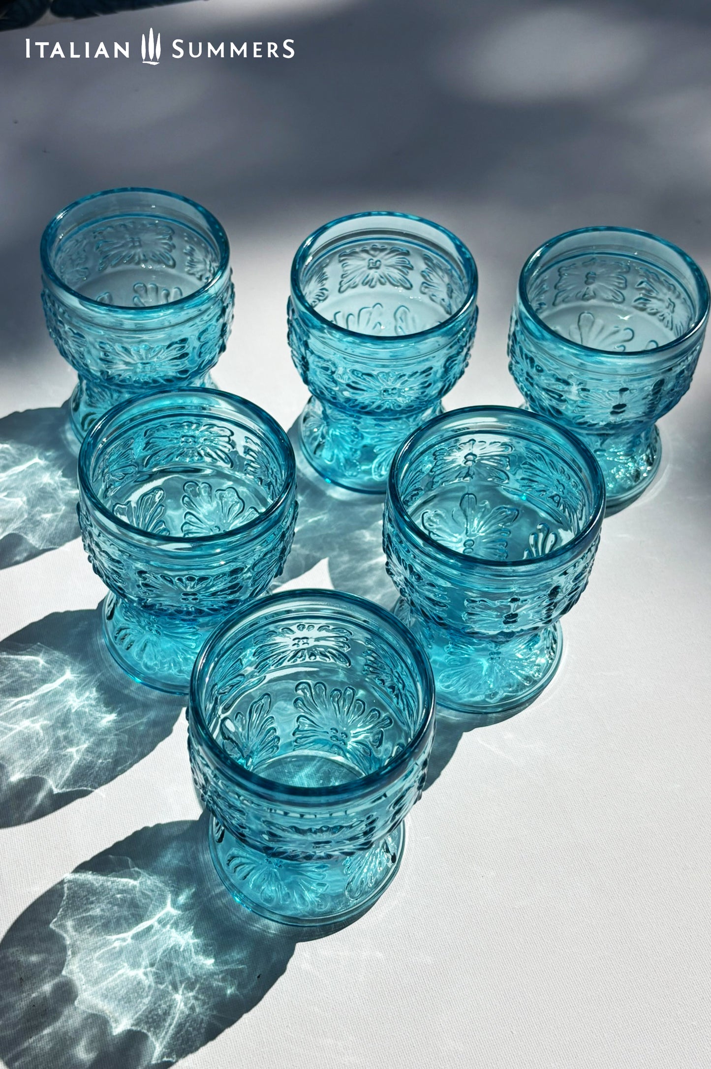 A blue glass carafe and six tumblers set, all embossed in flower patterns from the 1970s Italy, sits on a table with a colorful and bright tablecloth with blue floral patterns.