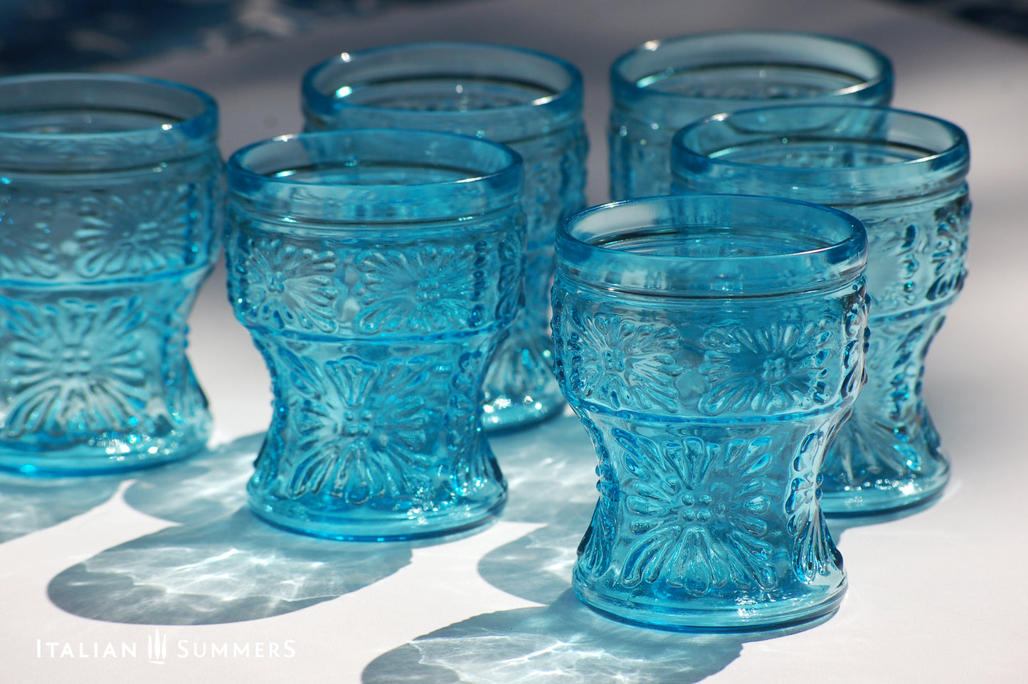 A blue glass carafe and six tumblers set, all embossed in flower patterns from the 1970s Italy, sits on a table with a colorful and bright tablecloth with blue floral patterns.