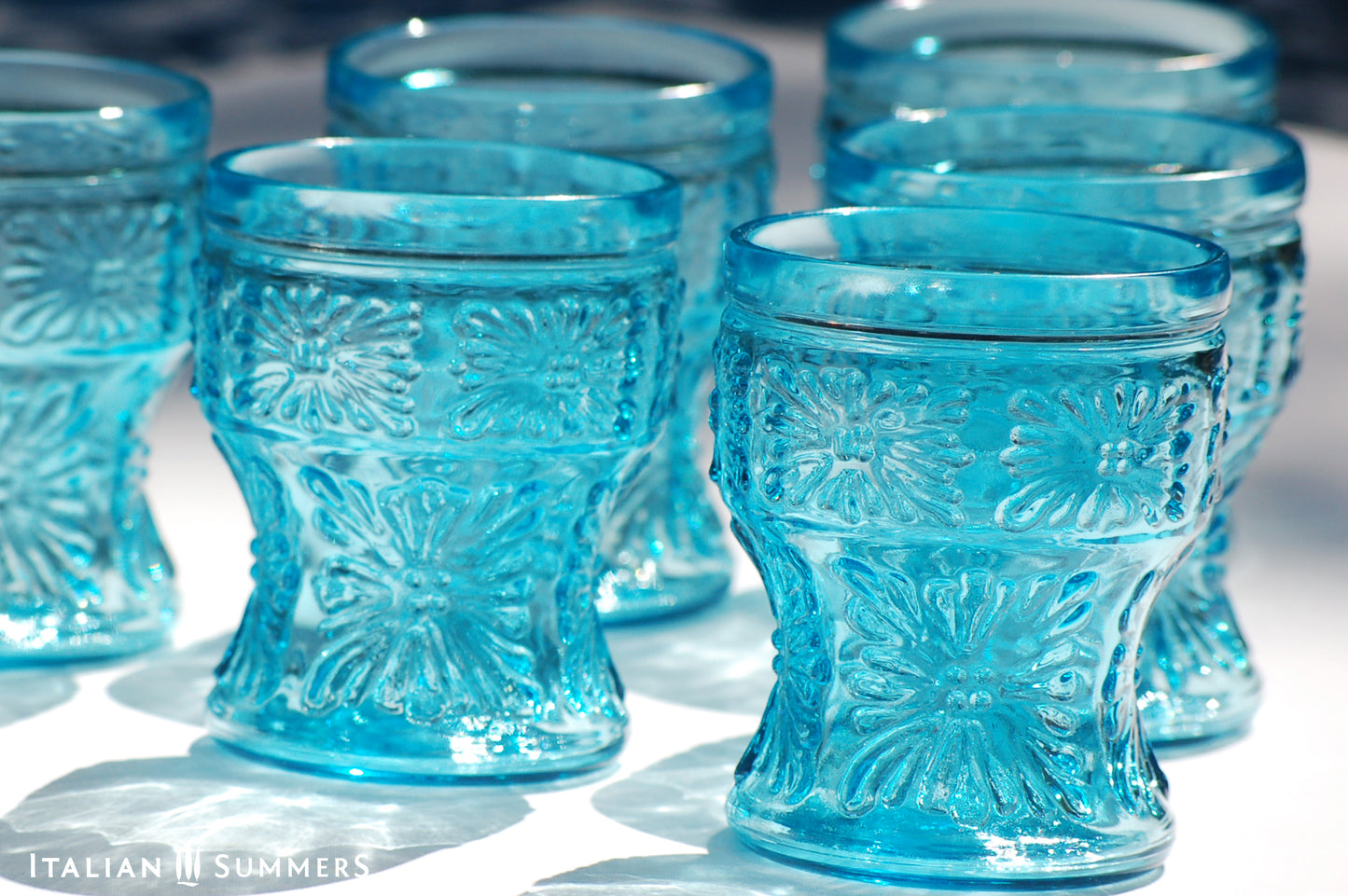 A blue glass carafe and six tumblers set, all embossed in flower patterns from the 1970s Italy, sits on a table with a colorful and bright tablecloth with blue floral patterns.