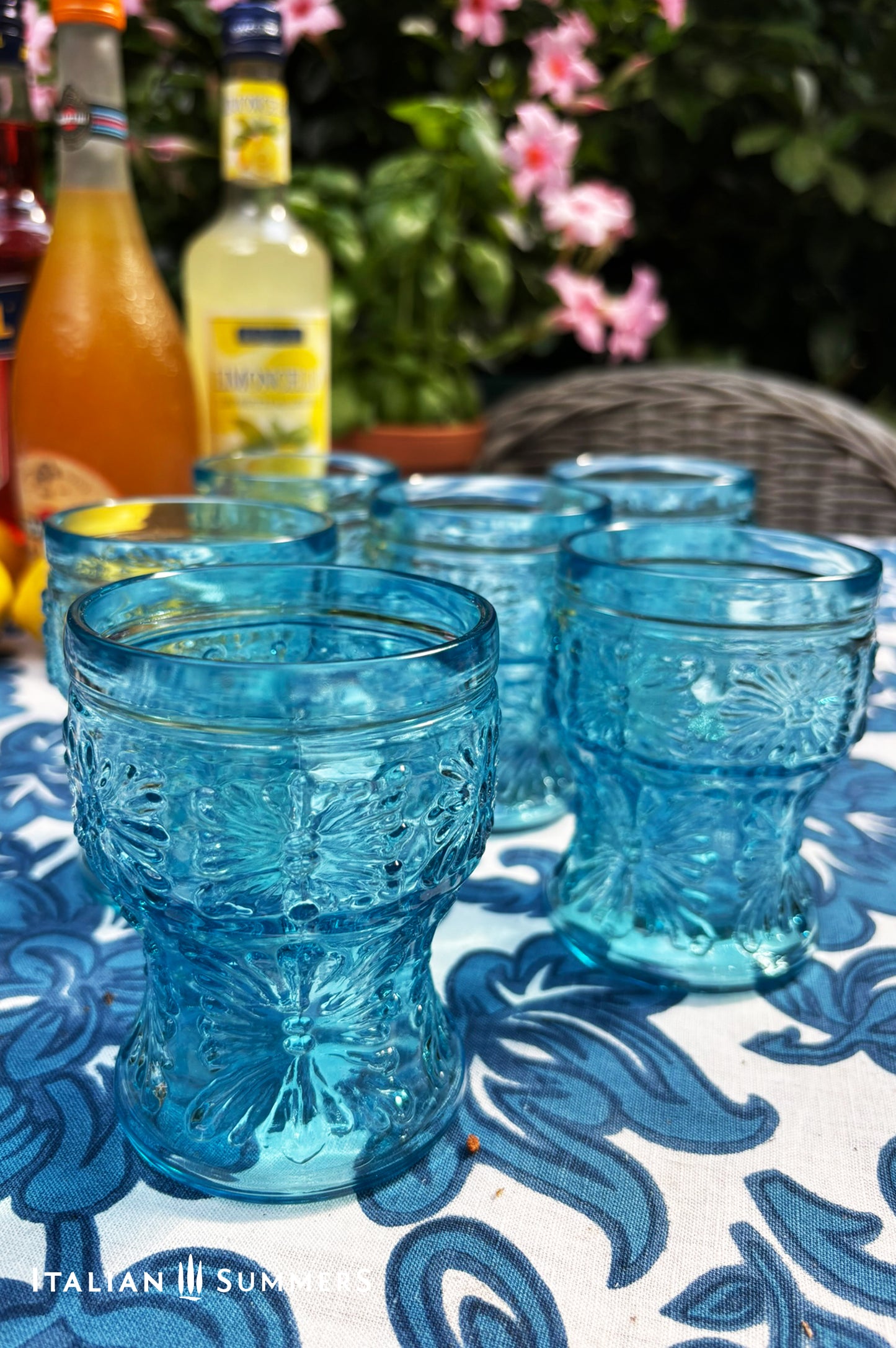 A blue glass carafe and six tumblers set, all embossed in flower patterns from the 1970s Italy, sits on a table with a colorful and bright tablecloth with blue floral patterns.