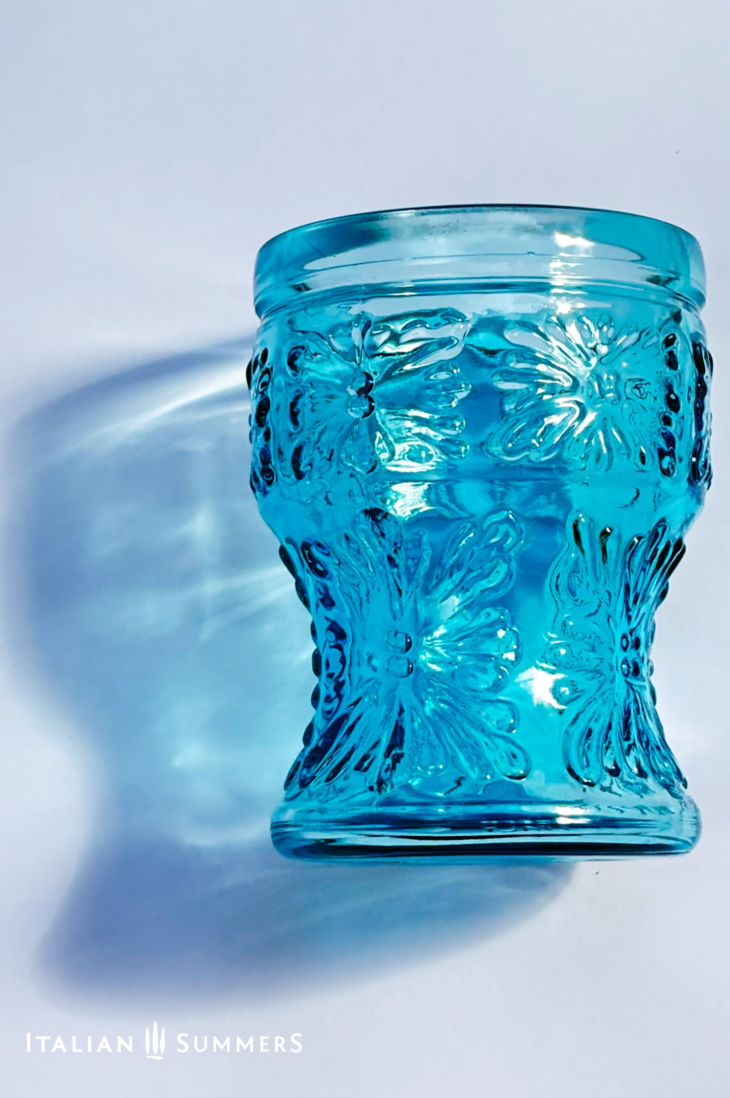 A blue glass tumbler embossed in flower patterns from the 1970s Italy, sits on a table with white tablecloth.