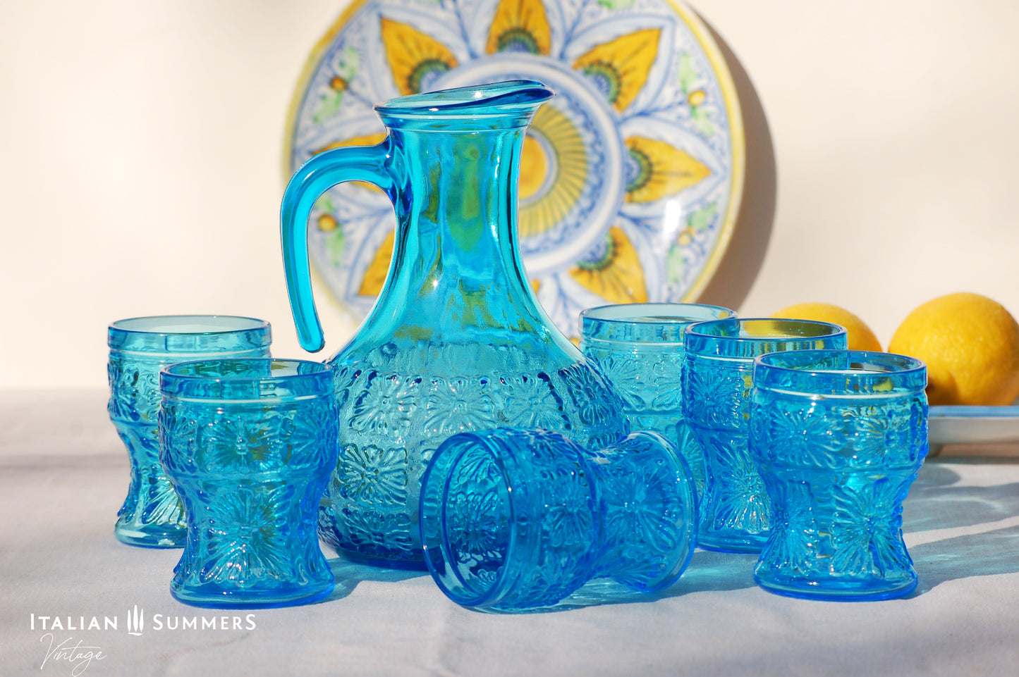 A blue glass carafe and six tumblers set, all embossed in flower patterns from the 1970s Italy, sits on a table with a colorful and bright tablecloth with blue floral patterns.