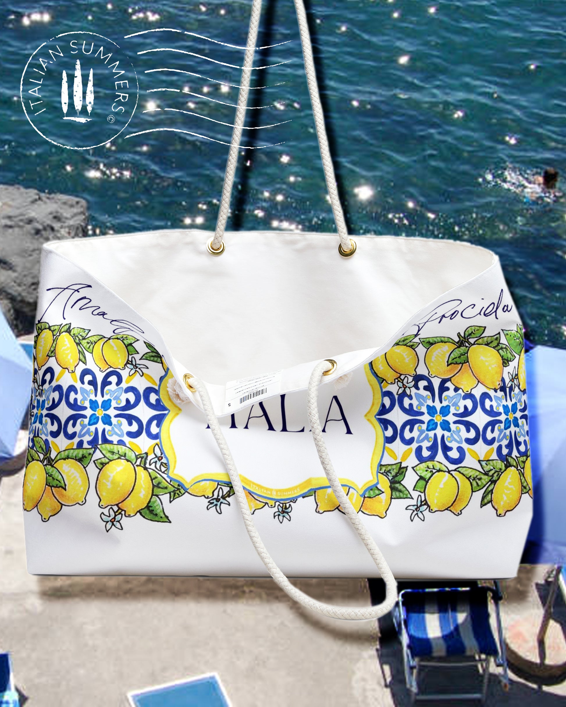 Italy-inspired XL Beach Bag  with printed blue Italian tiles in a  large band across the entire bag , framed with garlands of sunny, leafy Sorrento Lemons  with flowers. A large cartouche -like crest in the middle has the word  ITALIA - Seaside location names are writthen in flowing hand-script in blue across the top of the design. Sorrento, Positano, Ravello, Praiano, Capri, Amalfi, Procida,  made by Italian Summers