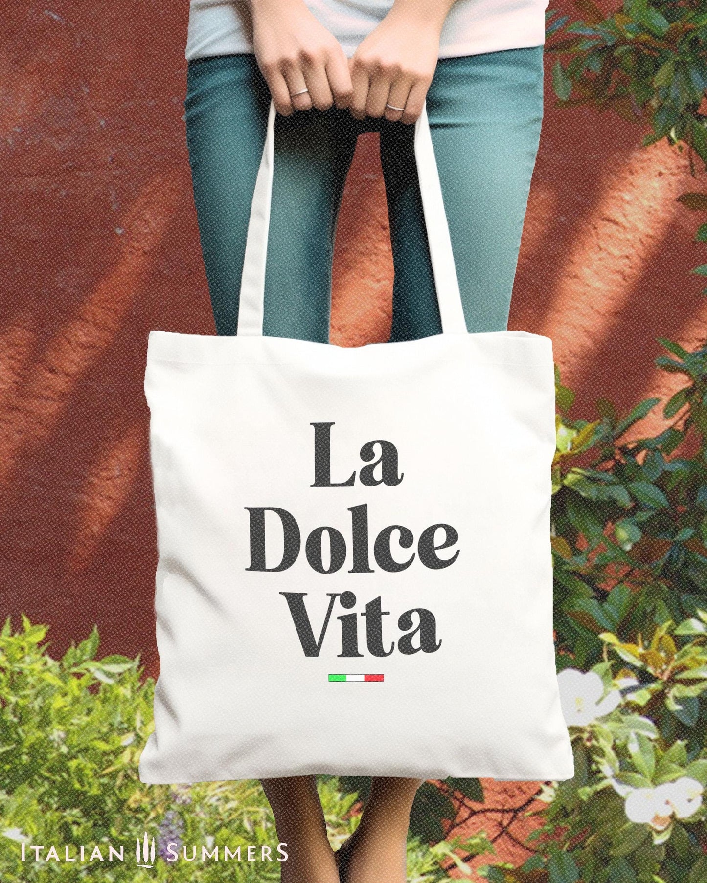 A cotton canvas tote bag with the text La Dolce Vita is held by a young woman in front of her legs. a small Italian flag decorates the bag.