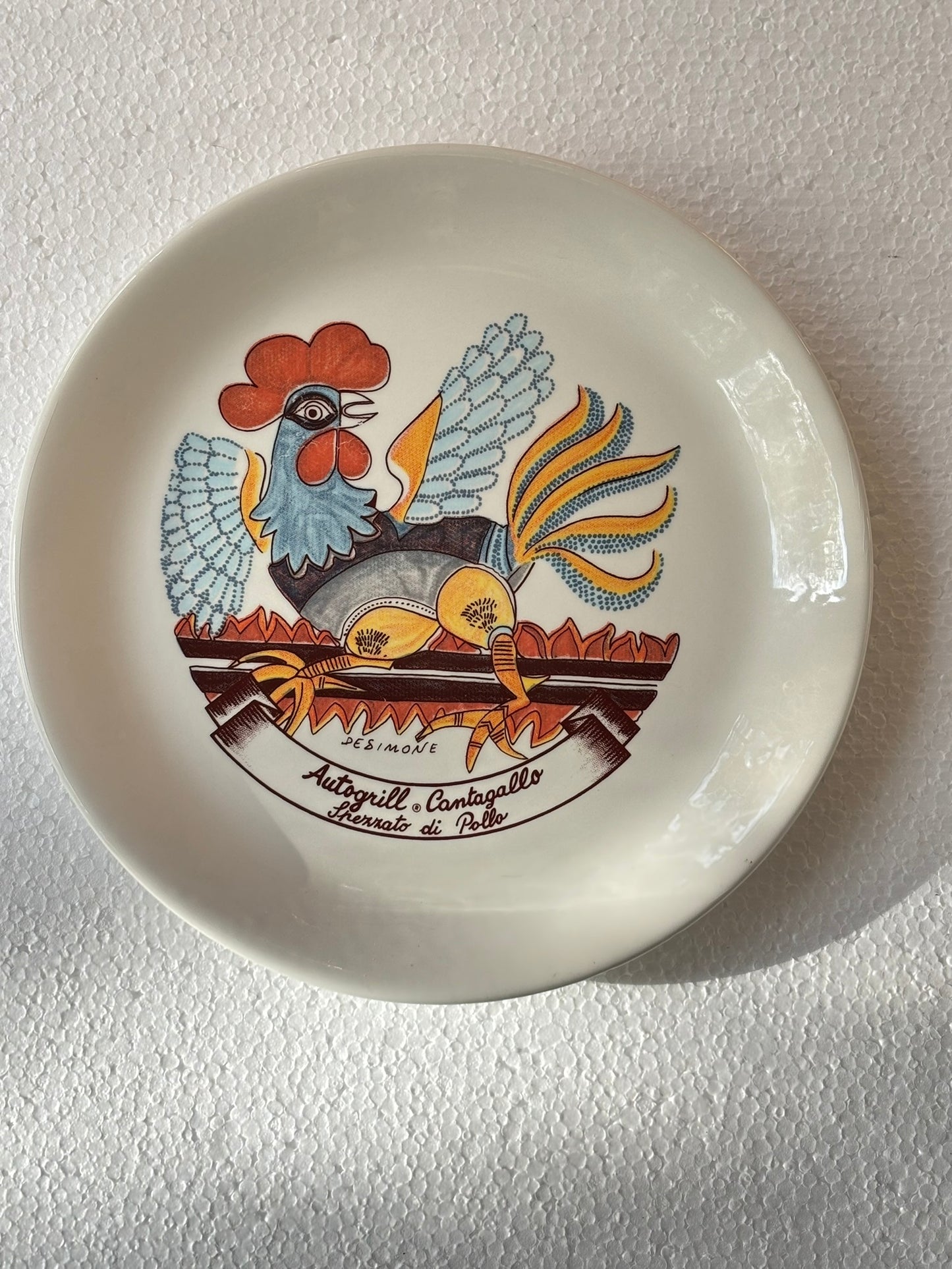 An Italian Vintage ceramic plate with the design by De Simone featuring a stylized rooster, with a cartouche of the name of an Autogrill restaurant and a typical dish of the same.