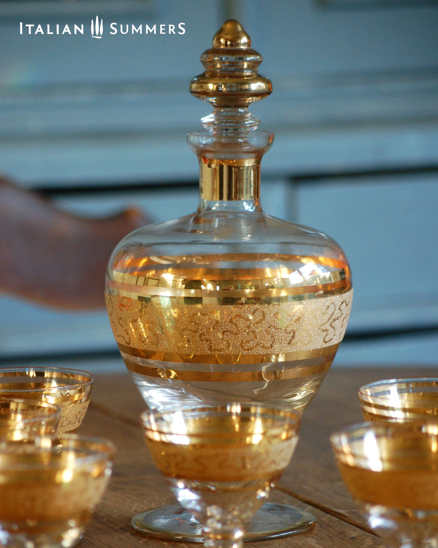 Decorative glass decanter with gold accents on a reflective surface, with 'Italian Summers' branding.