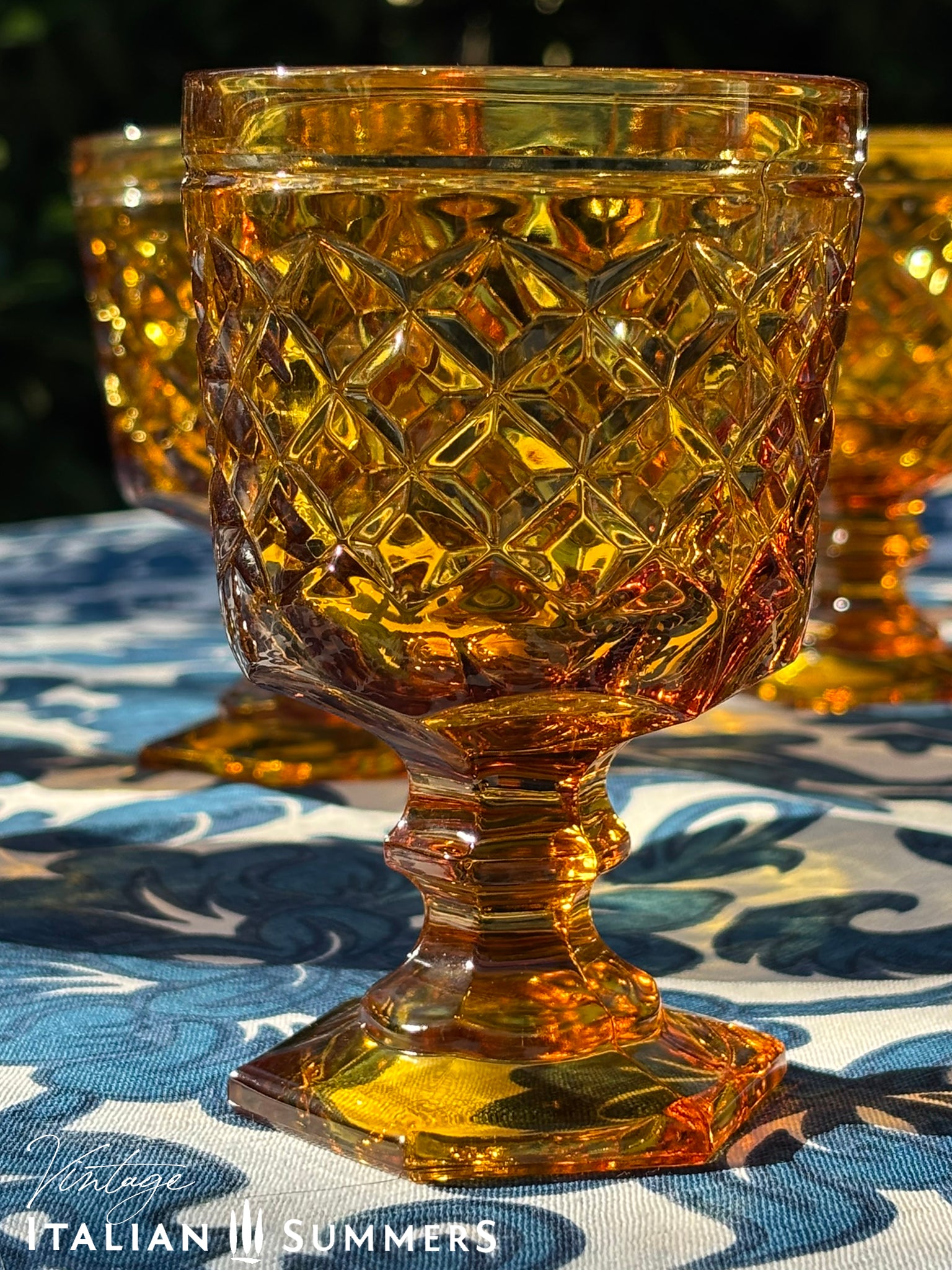 Set of Italian amber glass goblets and a carafe with the original Bormioli box on a floral patterned tablecloth.