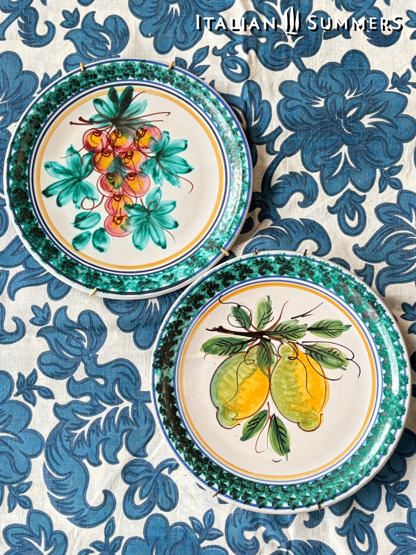 Two decorative Sicilian Caltagirone  plates with floral and fruit designs on a blue and white patterned fabric background.