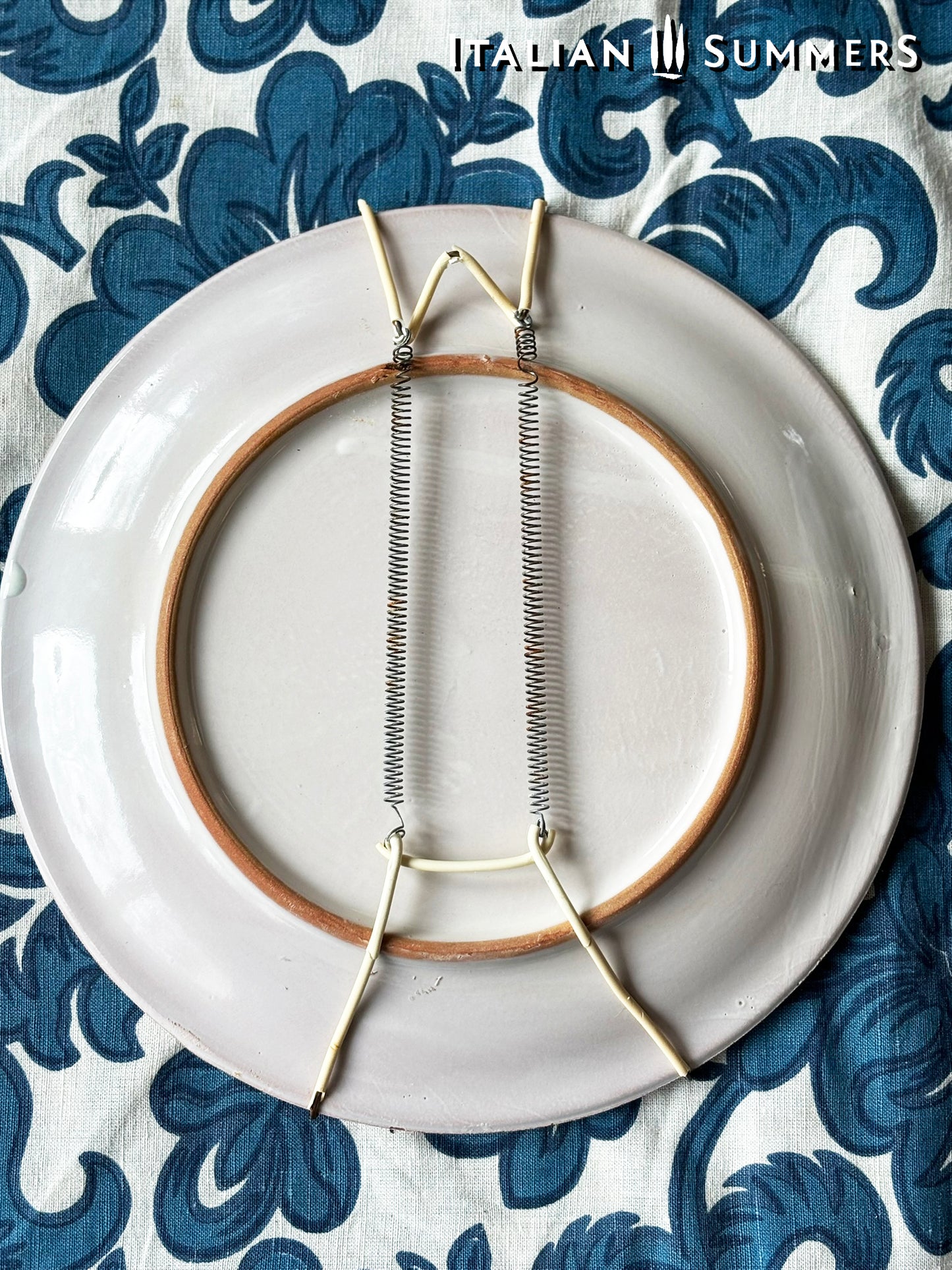 Backside of a decorative Sicilian Caltagirone  with hanging hook setup on a blue and white patterned fabric background.