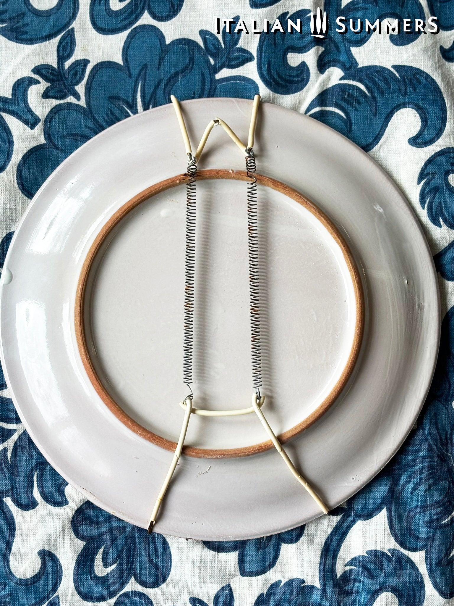 Backside of a decorative Sicilian Caltagirone  with hanging hook setup on a blue and white patterned fabric background.