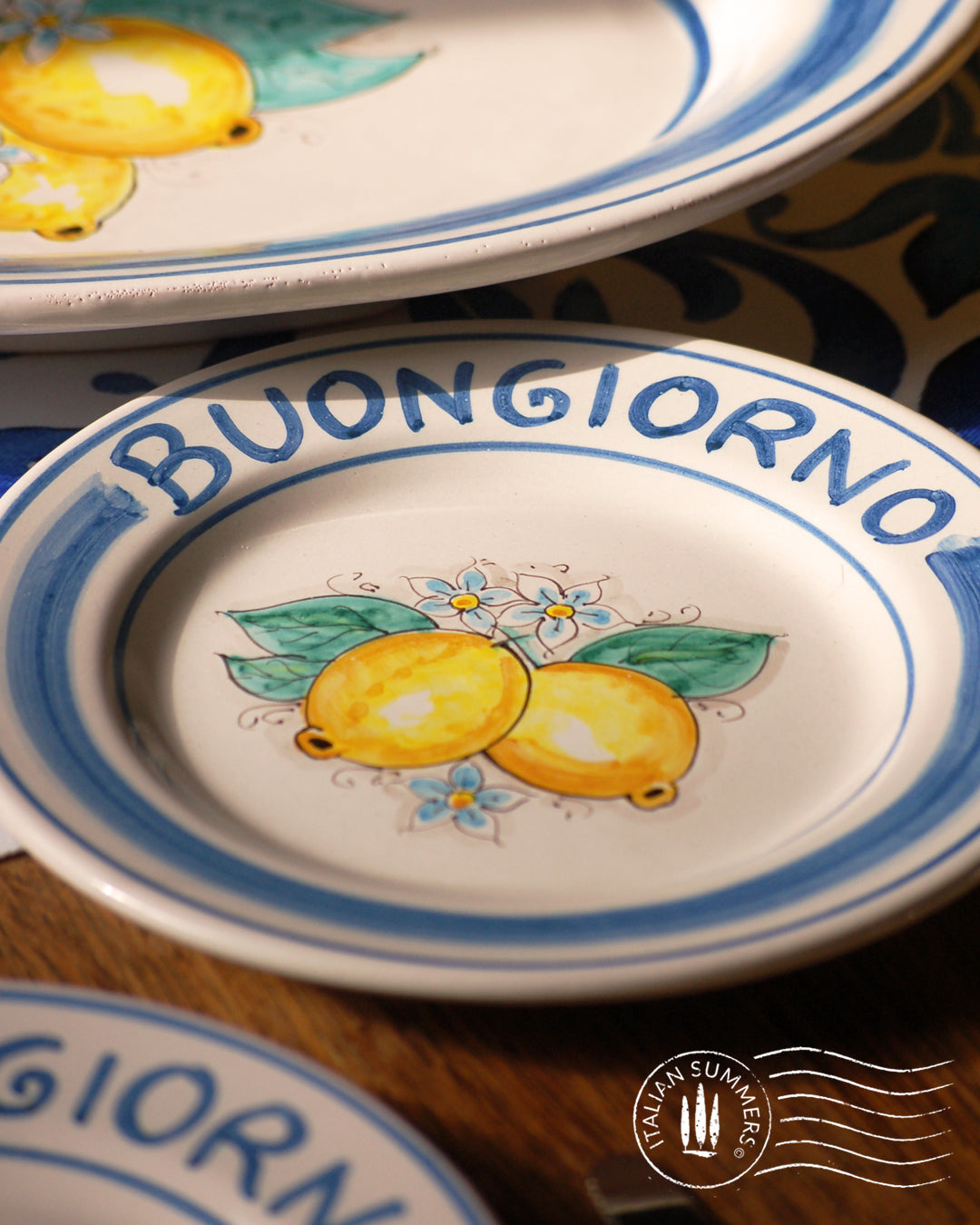 Ceramic plate Buongiorno Limoni handpainted in Sicily. This plate has the quote Biongiorno on the rim - handpainted - And colorfull lemons on the center of the plate with lemon flowers. On the rim of the plate there are blue paint stripes. The blue color is ultra marine. This plate is designed and sold by Italian Summers.