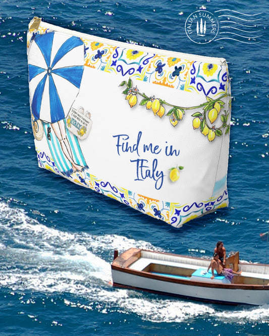 An Italy inspired white canvas clutch with the text Find me in Italy. On the front ther is a sketch of a lady on a striped  beach towel under a blue and white beach umbrella seen from above. On the top and bottom there is a rim with blue and yello Italian tiles. In the back there is a sketch of Positano and a Riva boat in the sea. Front and back have also some lemon branches. Designed by Italian Summers