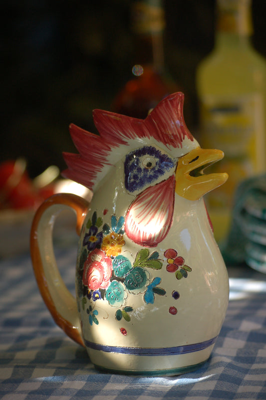 A Sicilian ceramic wine carafe featuring a vibrant rooster design with a floral pattern and a red flower, set against a checkered background.