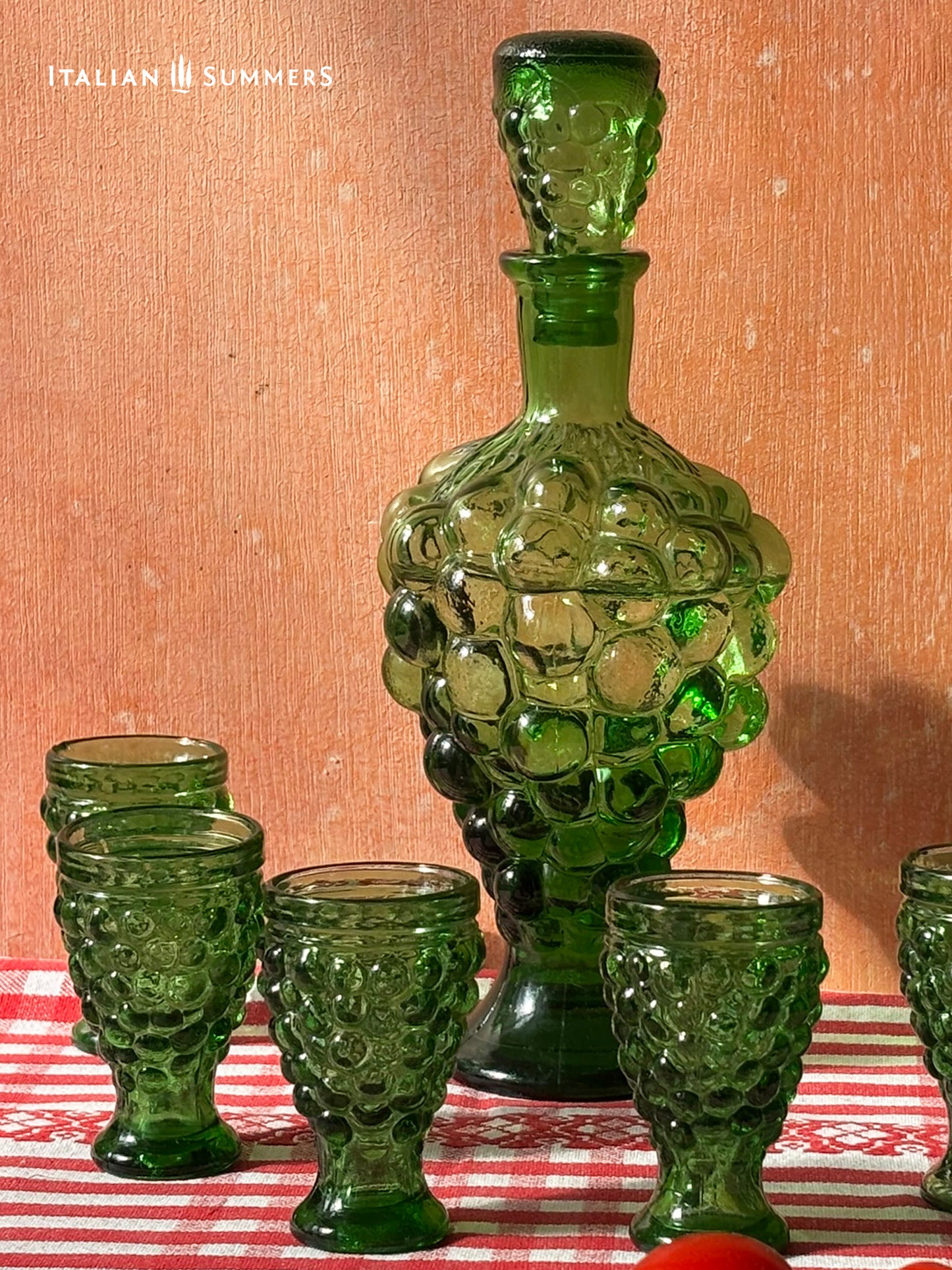 Green glass decanter and goblets on a red and white checkered tablecloth with 'Italian Summers' branding.