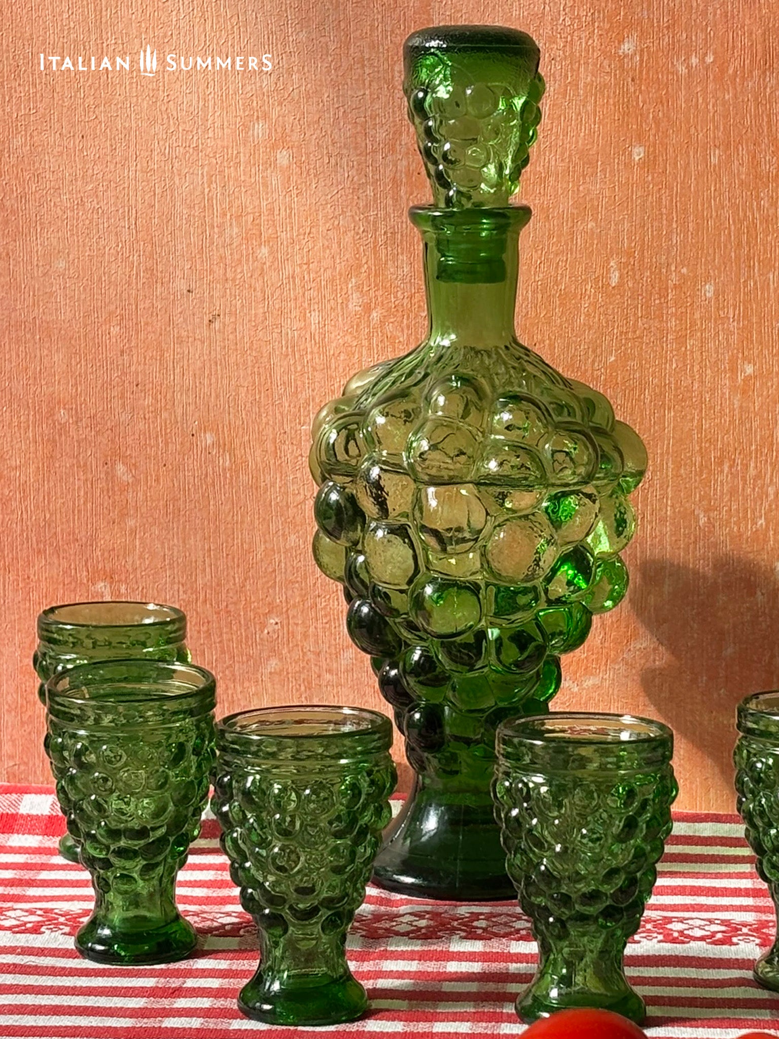 Green glass decanter and goblets on a red and white checkered tablecloth with 'Italian Summers' branding.