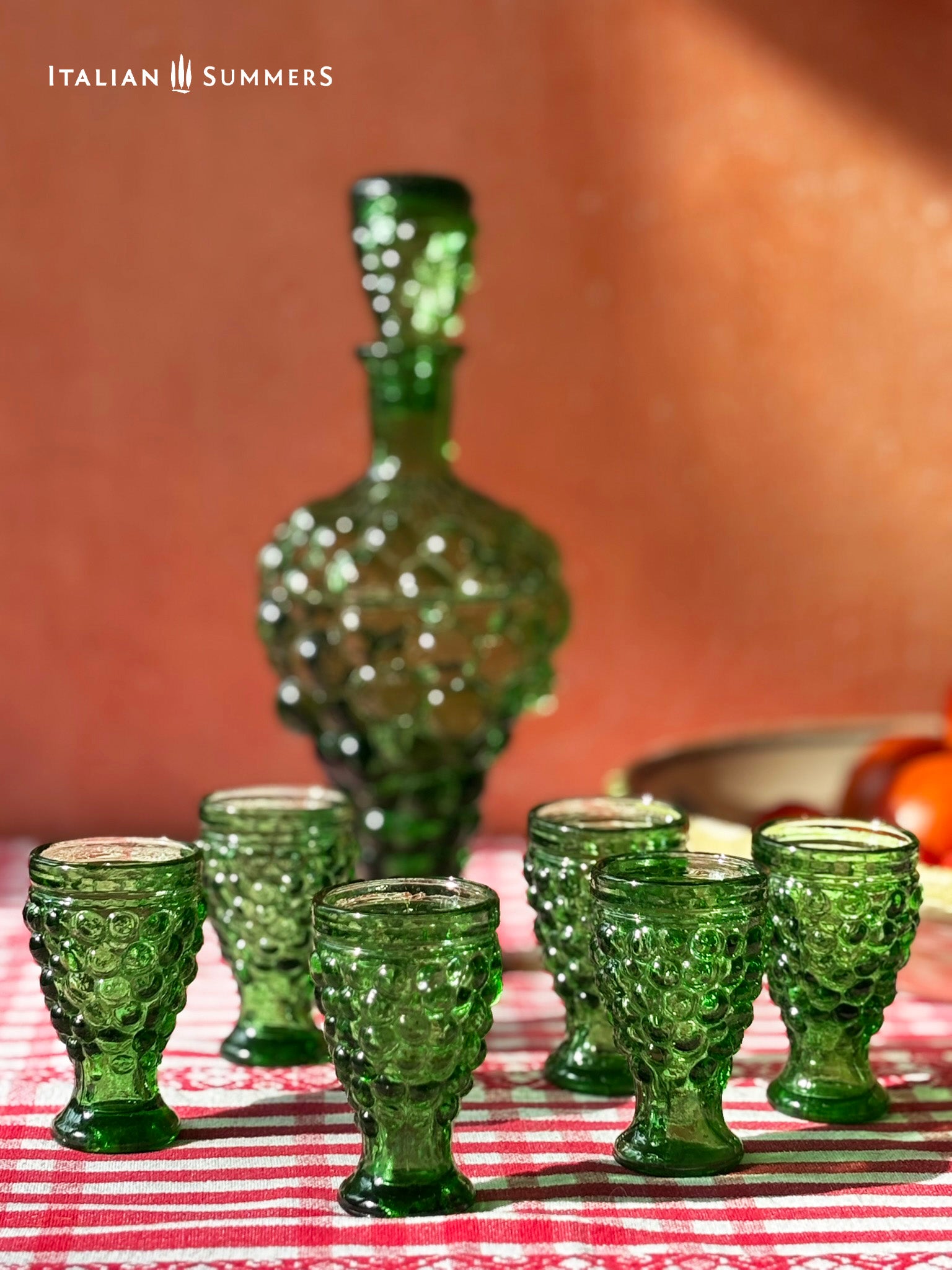 Set of green glass goblets and a decanter on a red and white checkered tablecloth with 'Italian Summers' branding.