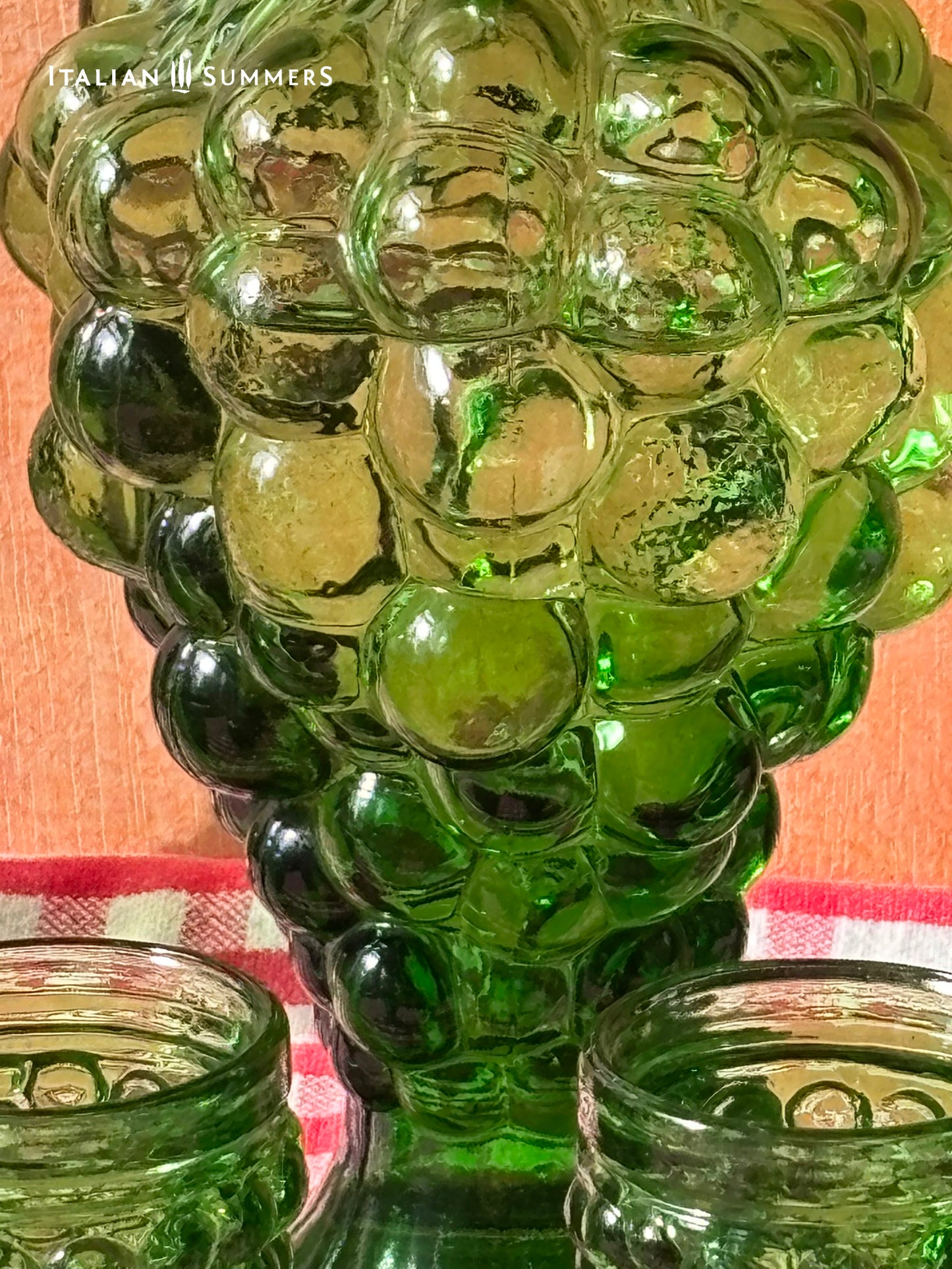 Green glass embossed grape bunch shaped decanter and goblets on a checkered tablecloth with 'Italian Summers' branding.