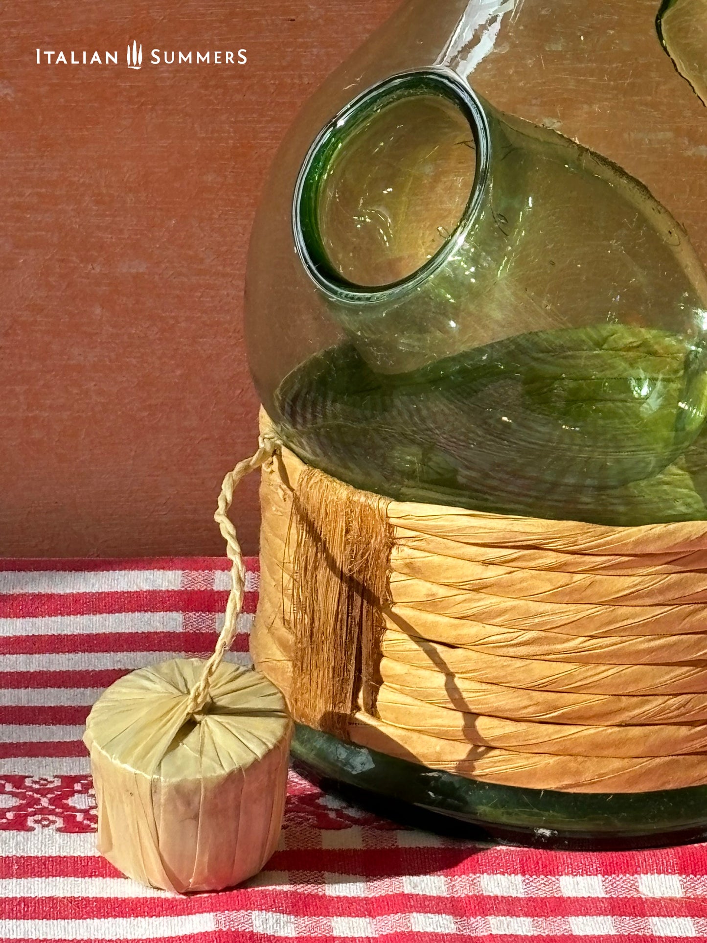 detail of an Empoli glass bottle with natural wrap and small ice container built-in, on a red and white checkered tablecloth, with 'Italian Summers' branding.