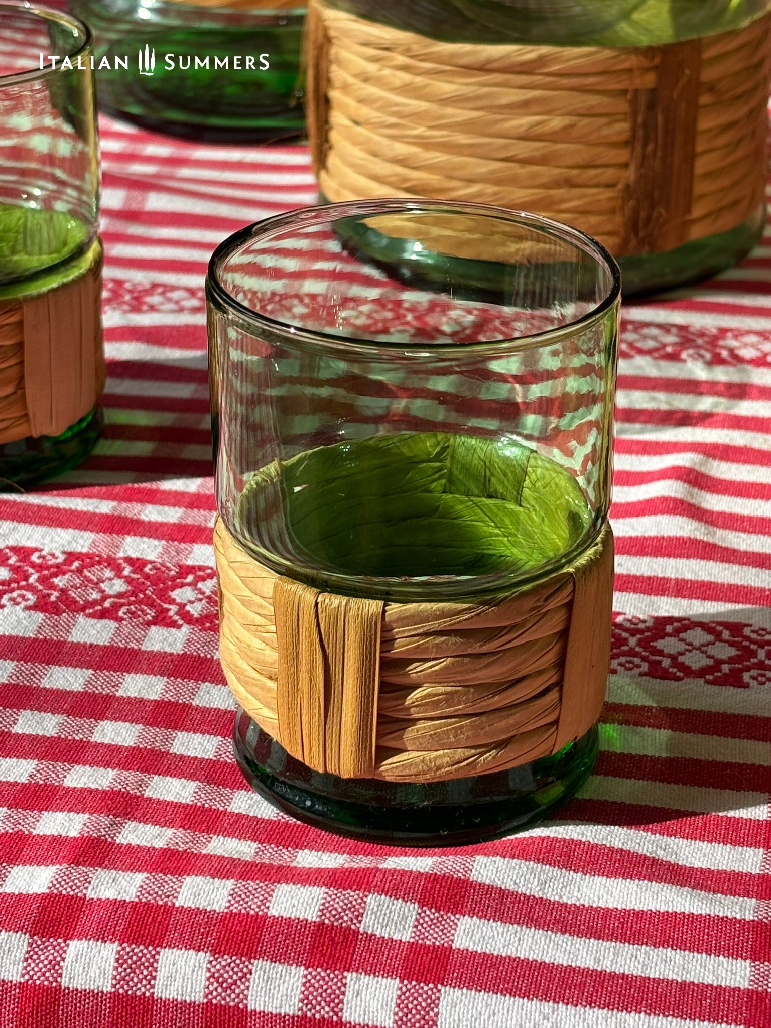 Glass with woven wicker sleeve on a red and white checkered tablecloth, brand 'Italian Summers' visible.