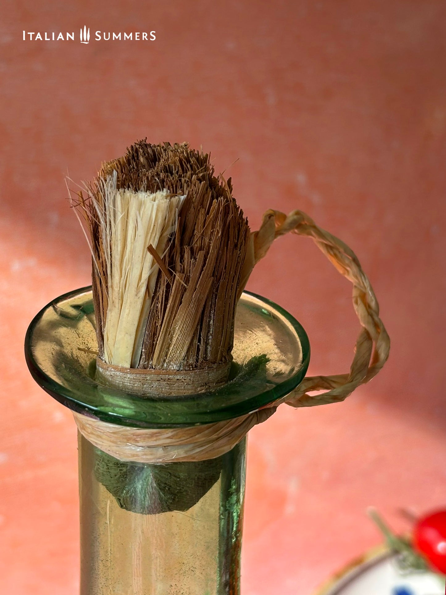 detail of he straw stopper on a Green Empoli vintage glassware and decanter on a table with a checkered tablecloth, tomatoes, and 'Italian Summers' branding.