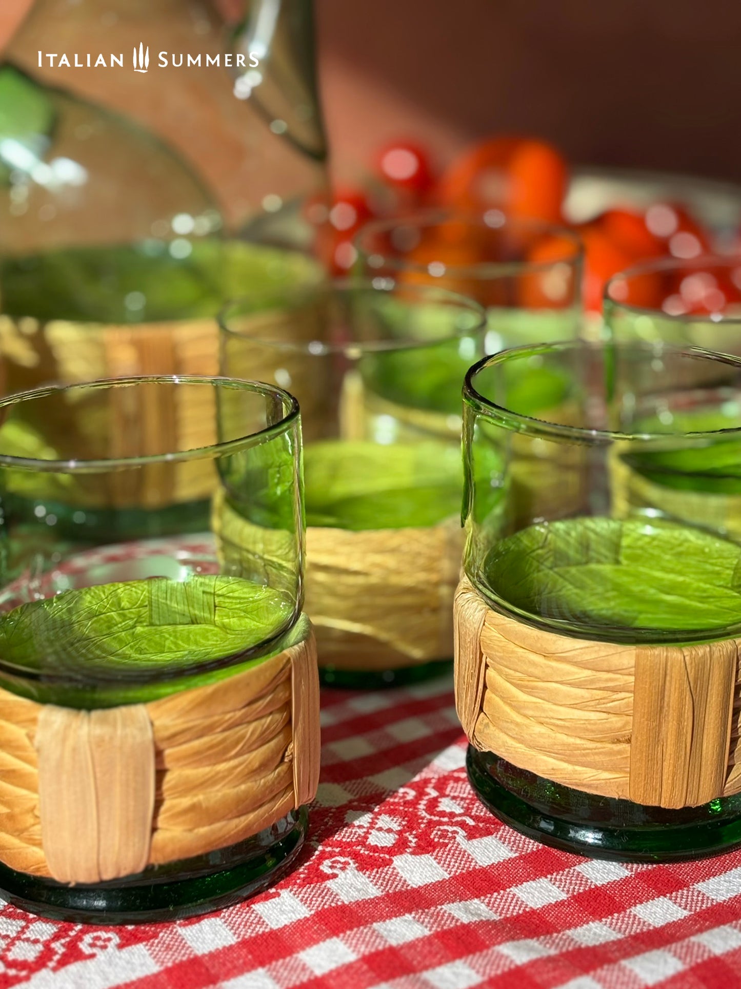 Green glasses with woven  straw sleeves on a red and white checkered tablecloth, featuring 'Italian Summers' branding.