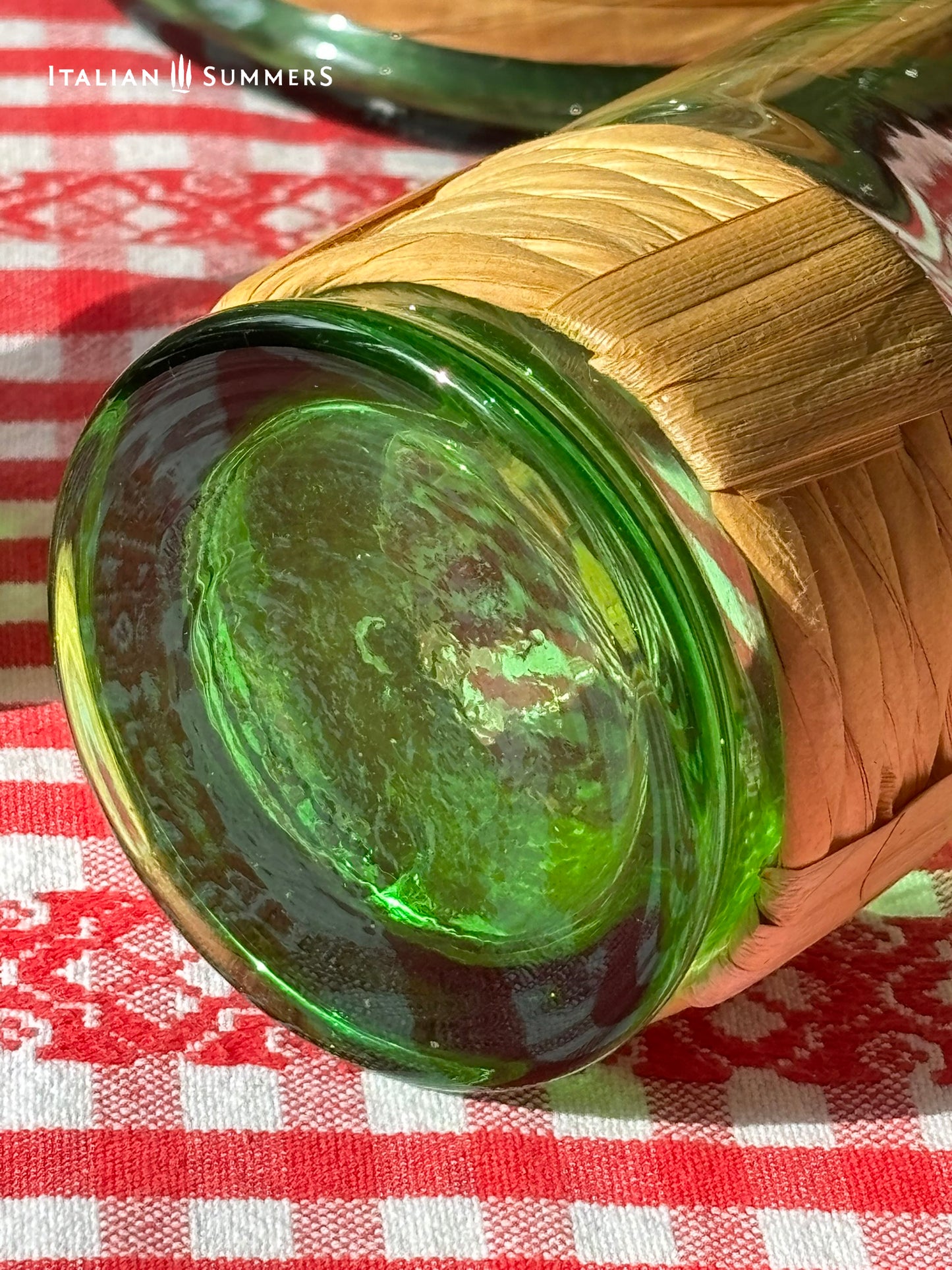detail of the bottom of a wine glass from a Green Empoli vintage glassware and decanter set on a table with a checkered tablecloth, tomatoes, and 'Italian Summers' branding.