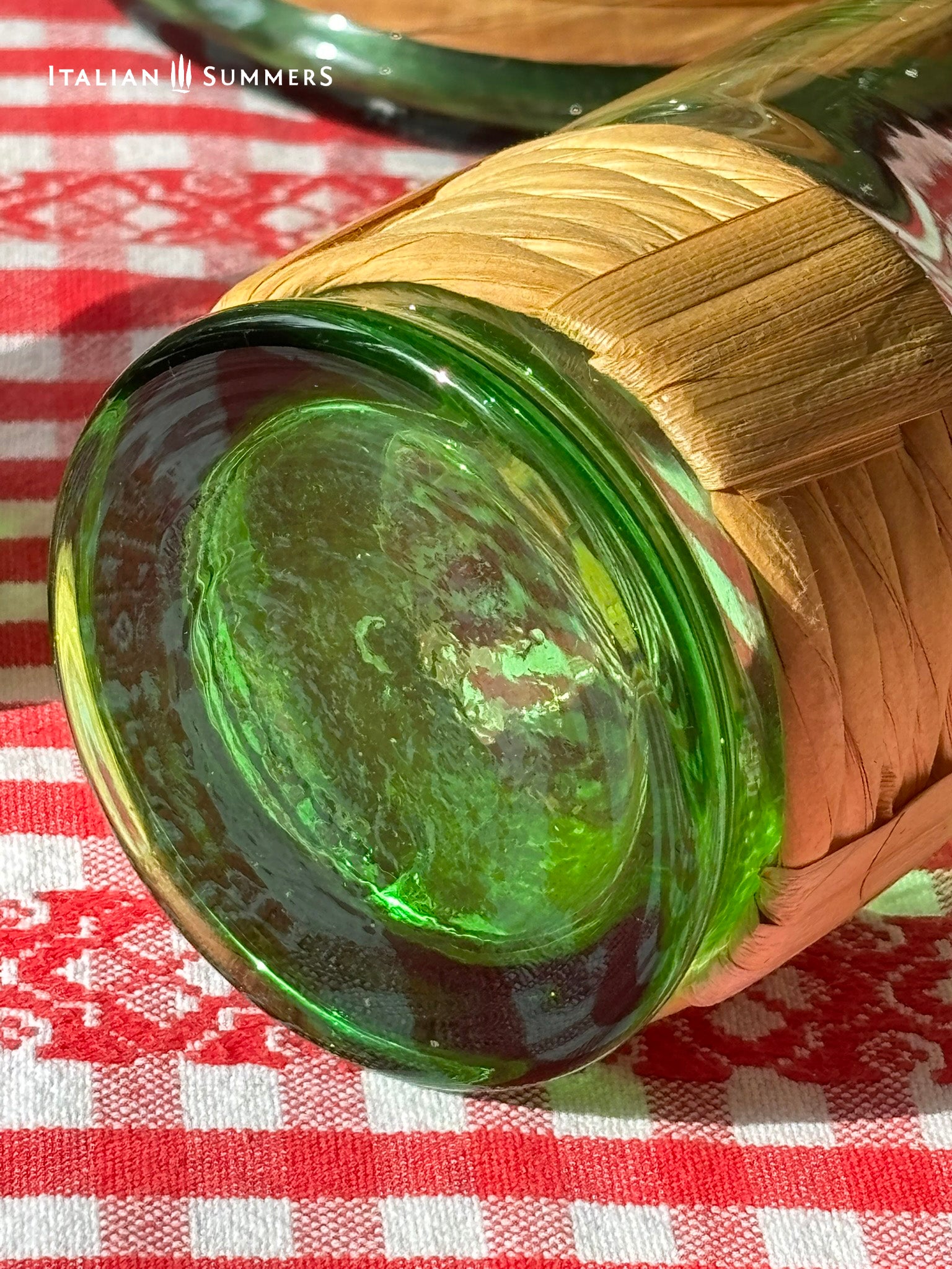 detail of the bottom of a wine glass from a Green Empoli vintage glassware and decanter set on a table with a checkered tablecloth, tomatoes, and 'Italian Summers' branding.