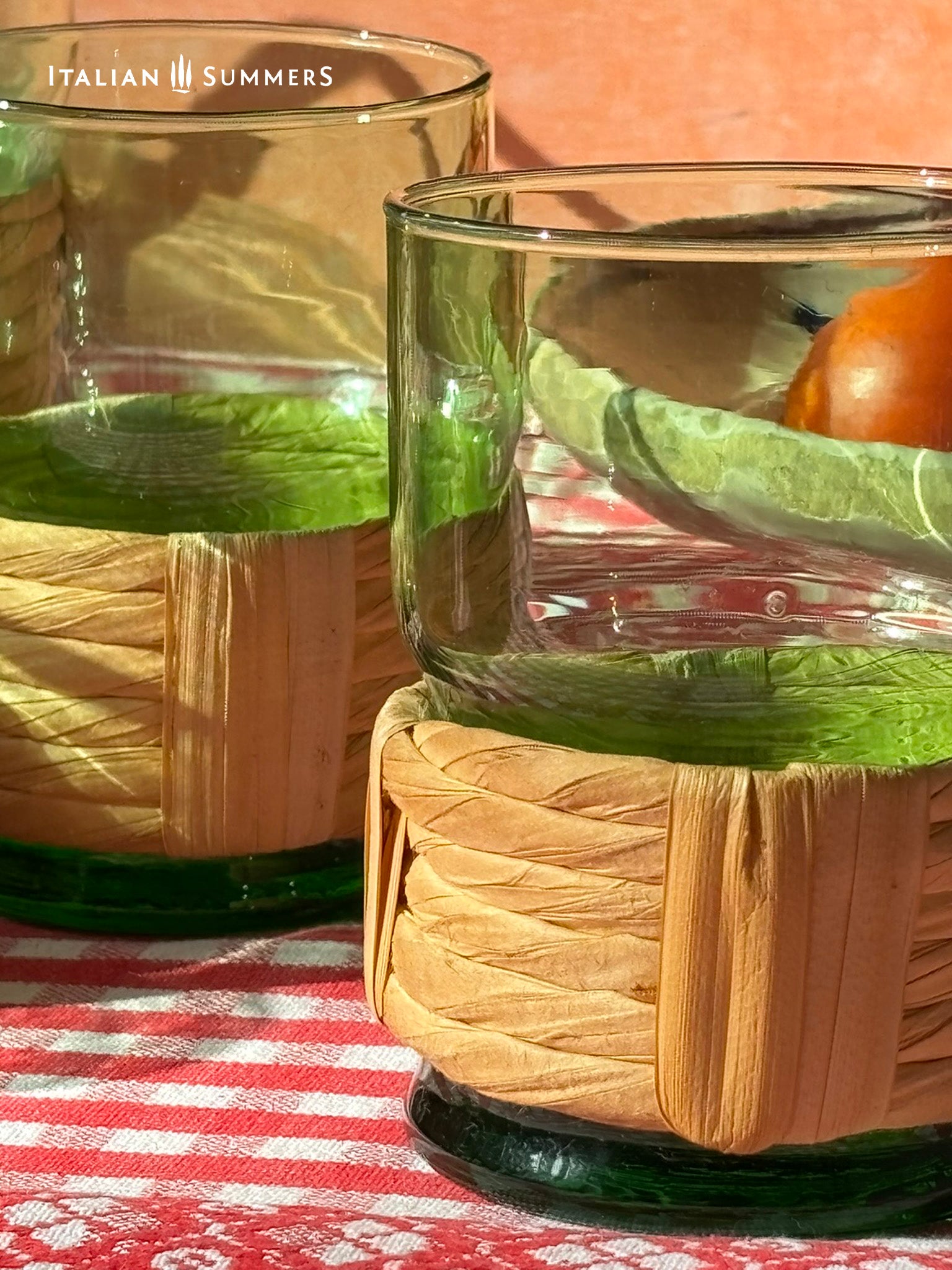Green Empoli vintage wine glasses with straw woven sleeves, part of a  decanter and six glass set on a table with a checkered tablecloth, tomatoes, and 'Italian Summers' branding.