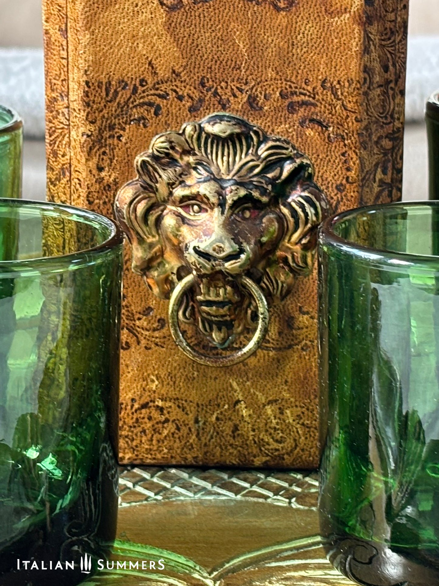 Decorative leather-covered whiskey decanter with lion head knocker and green glasses on a textured surface