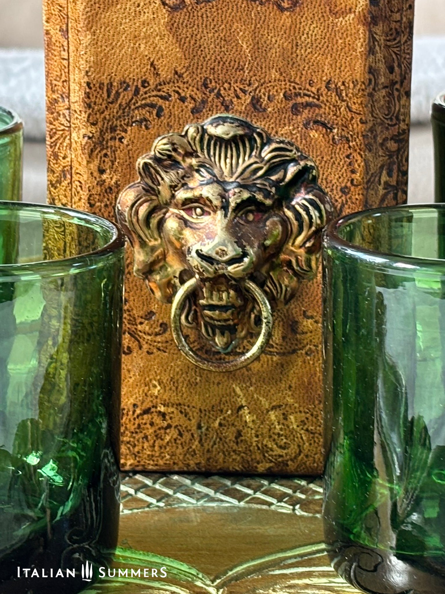 Decorative leather-covered whiskey decanter with lion head knocker and green glasses on a textured surface
