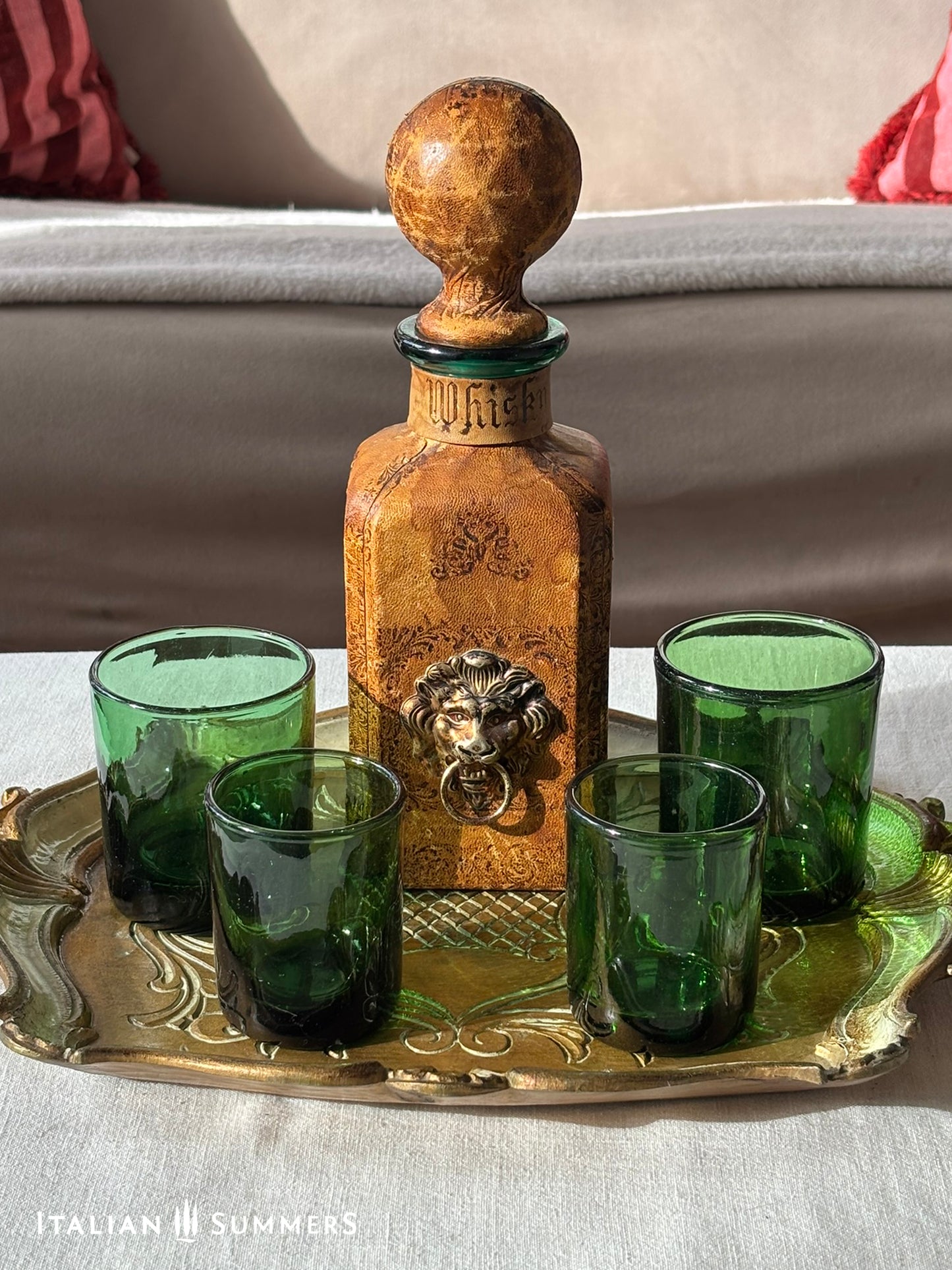 vintage leather-covered decanter with a lion head, round stopper and green glass tumblers on a gold-colored tray.