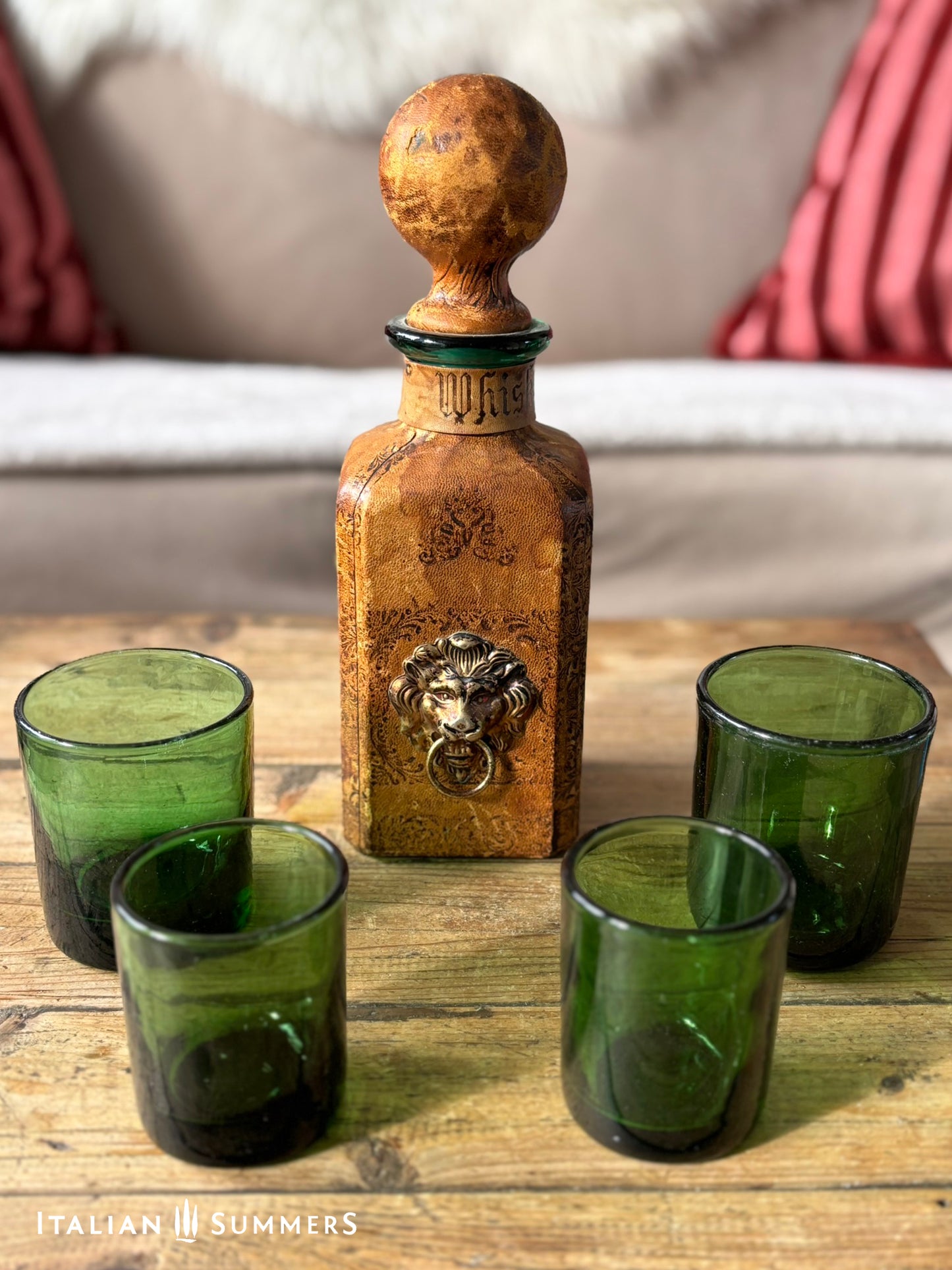 leather-covered whiskey decanter with a lion emblem and four green glasses on a wooden surface.