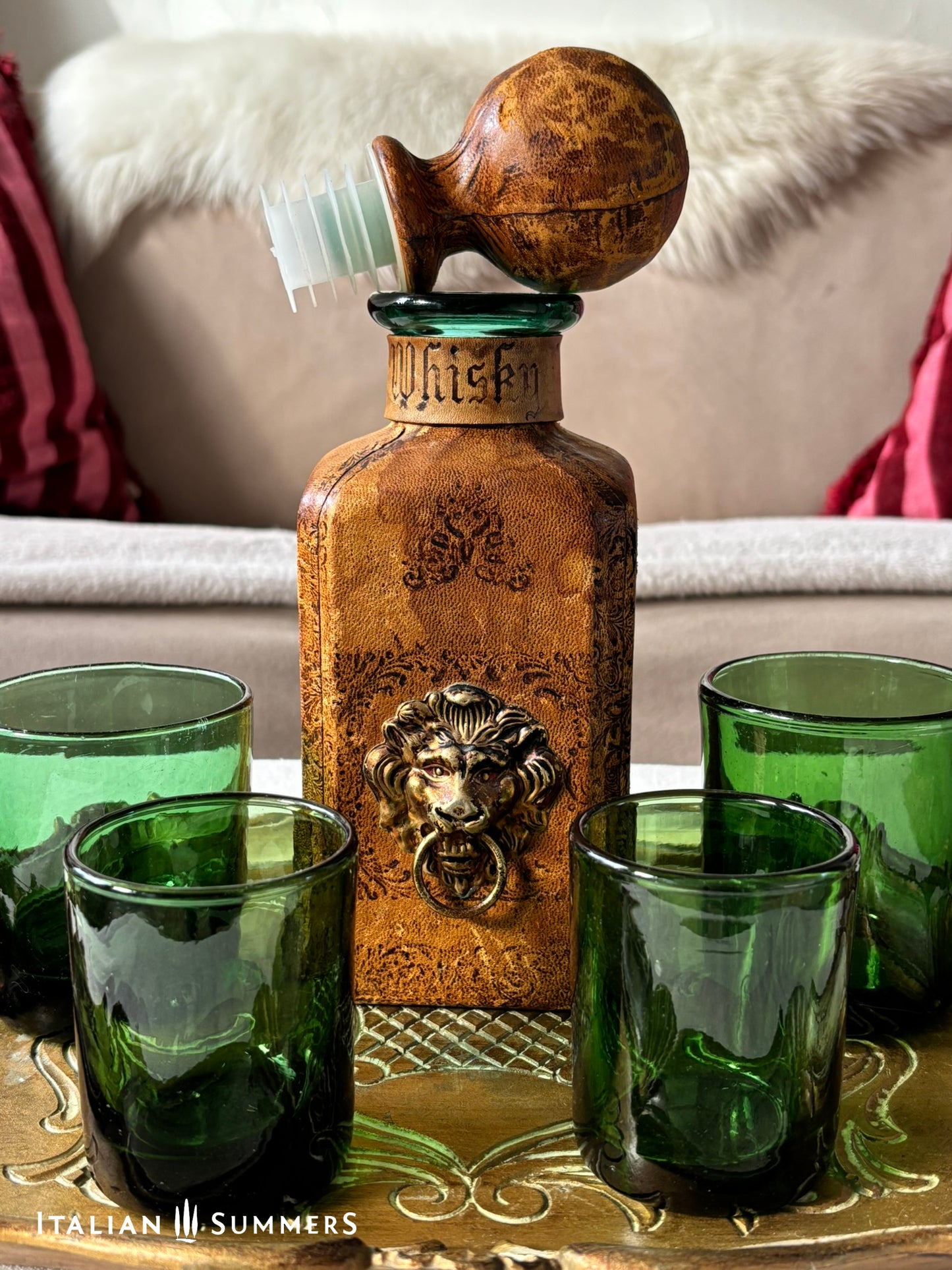 leather-covered whiskey decanter with lion emblem and green glasses on a decorative tray, brand 'Italian Summers' visible.
