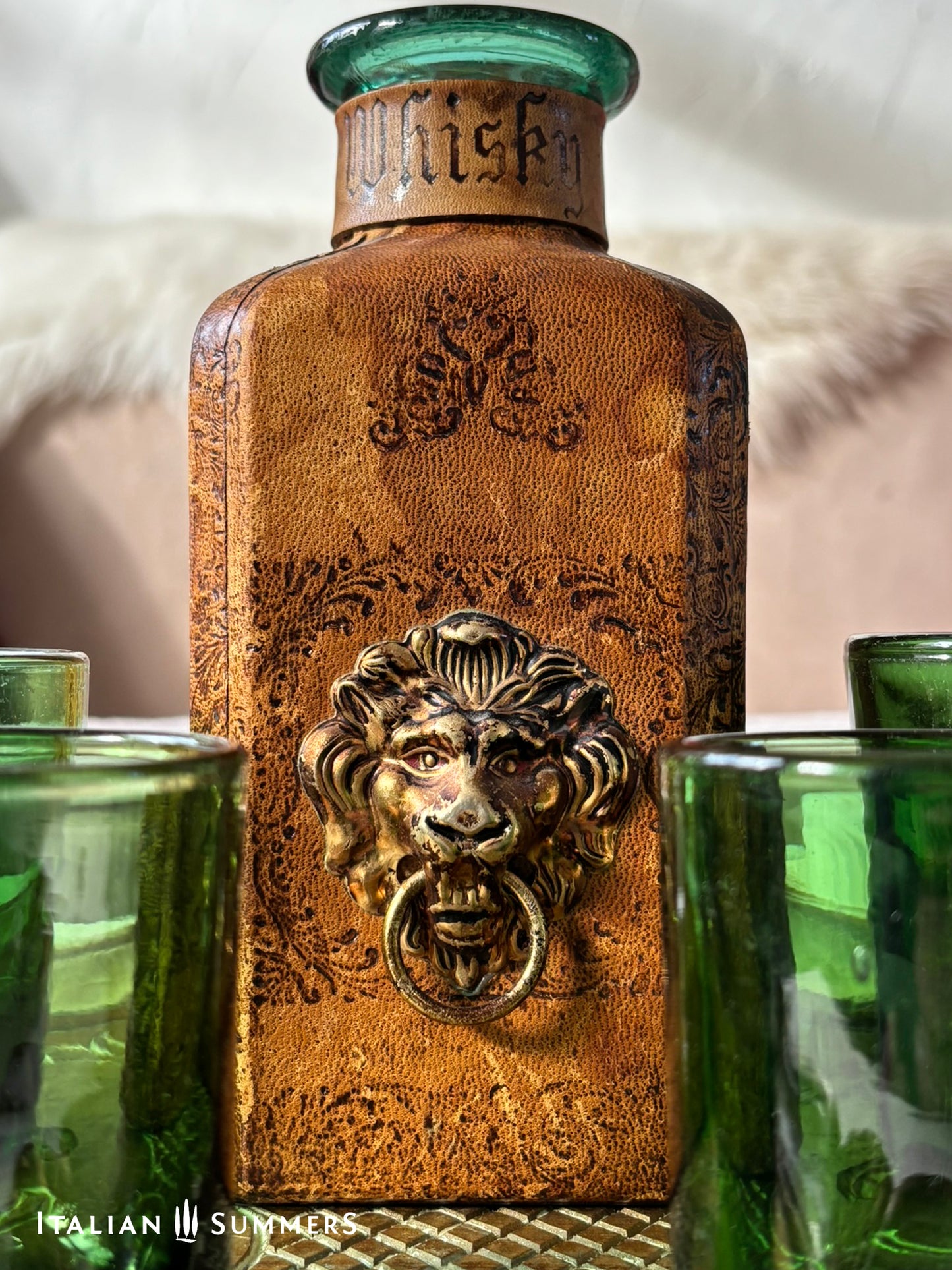 Decorative whiskey bottle with lion head knocker and green glasses on a textured surface.