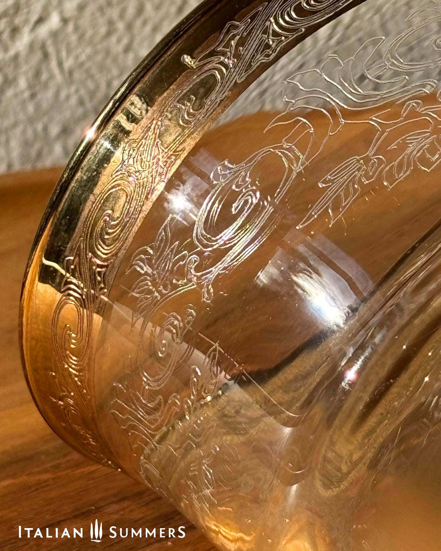 Glass coupe with decorated gold rim featuring intricate  floral filigree designs on a wooden surface, brand 'Italian Summers' visible.